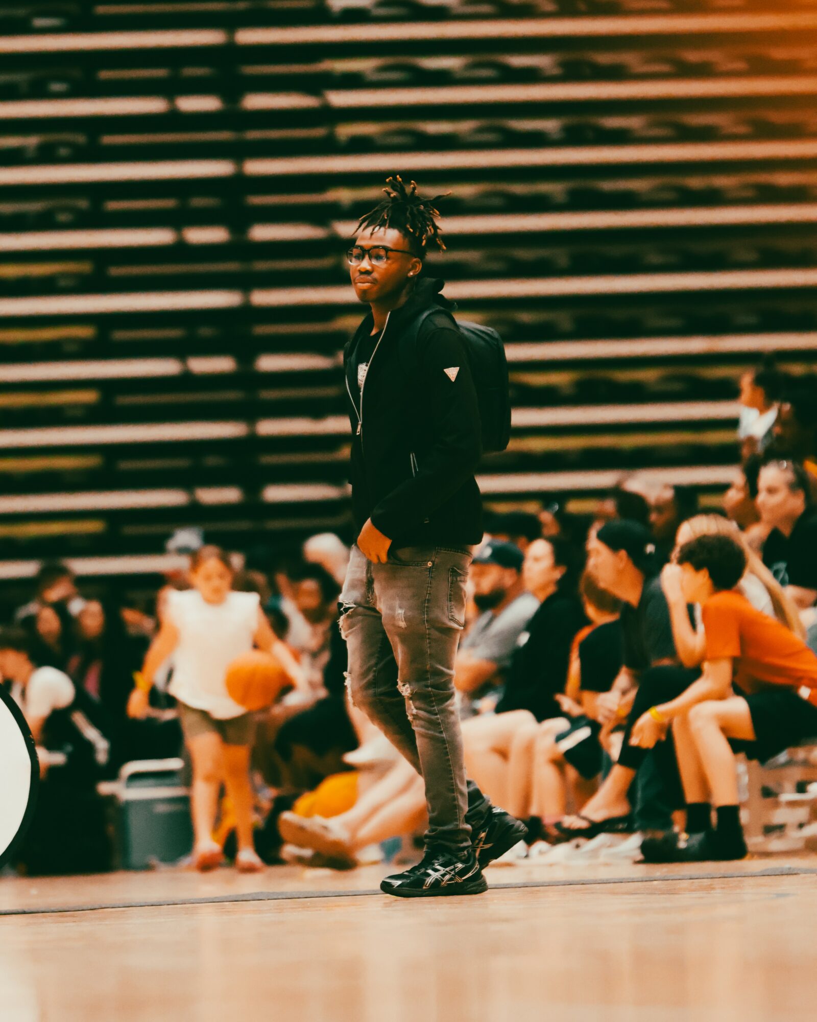 Person standing on a basketball court with spectators seated behind, wearing glasses, a black hoodie, and jeans.