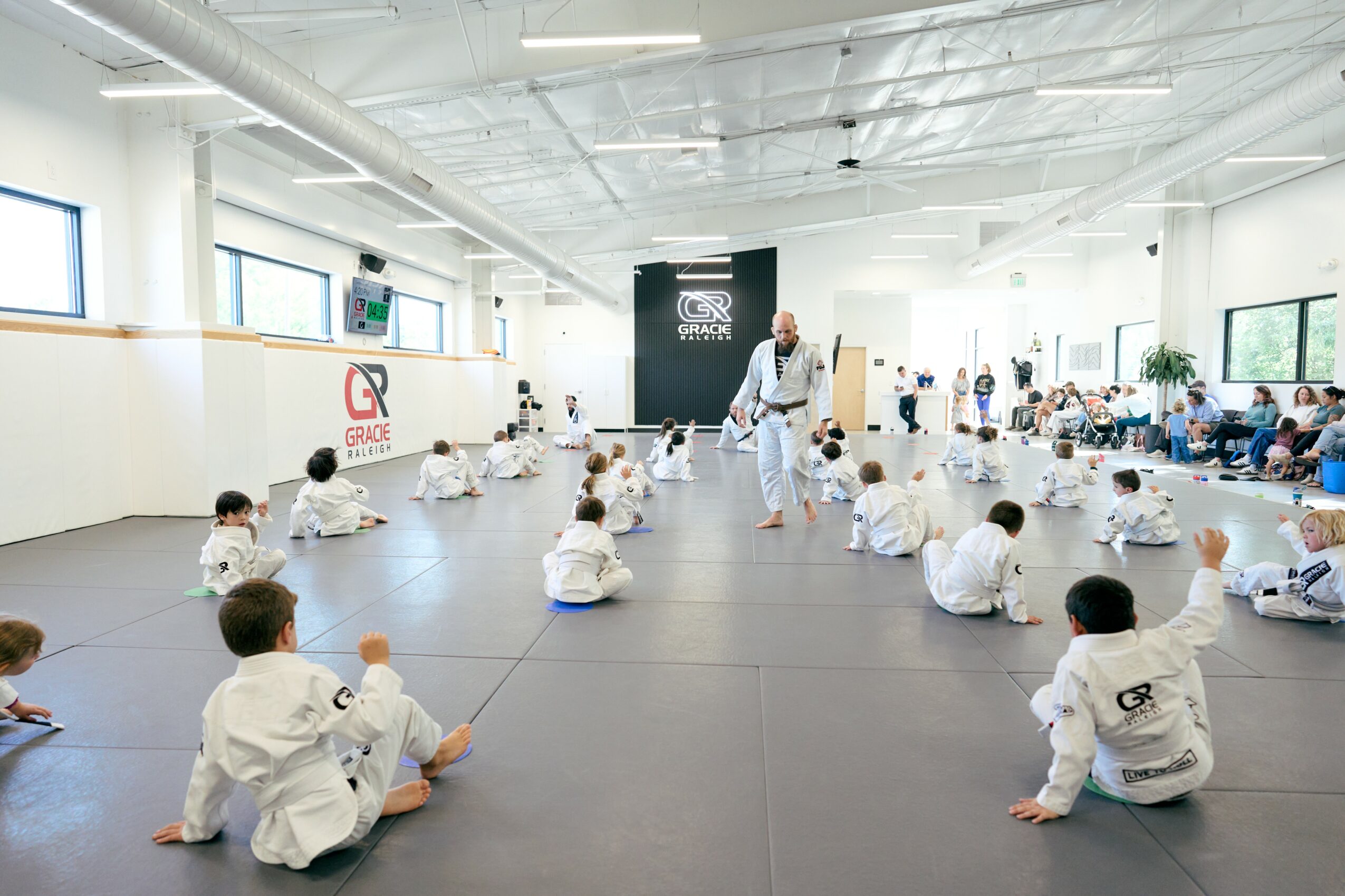Martial arts class with children sitting on mats, instructor demonstrating techniques, in a bright, spacious room with large windows.