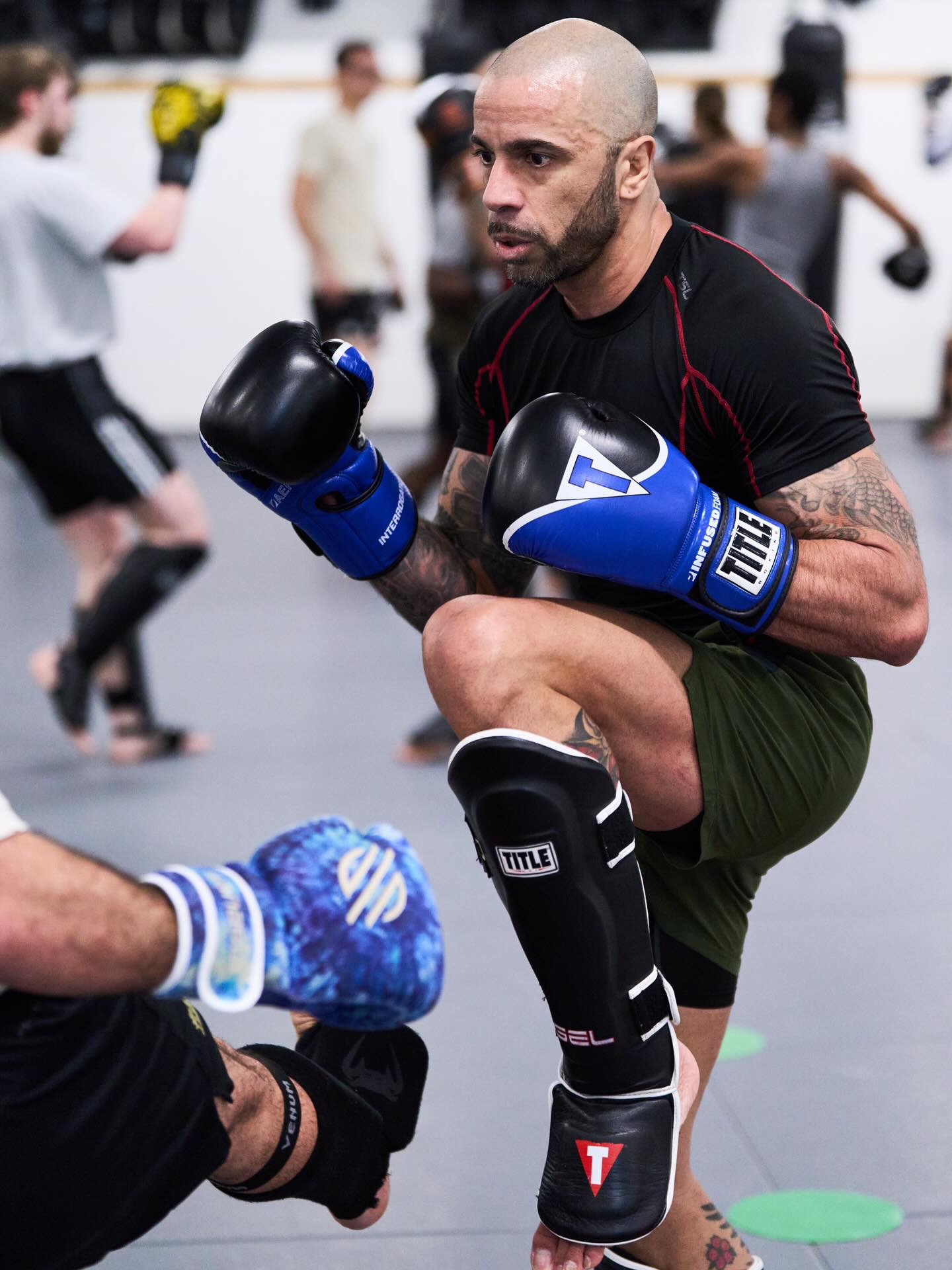 Bald man in black shirt and green shorts practicing boxing with gloves, kneeling on one knee in a gym, others training in background.