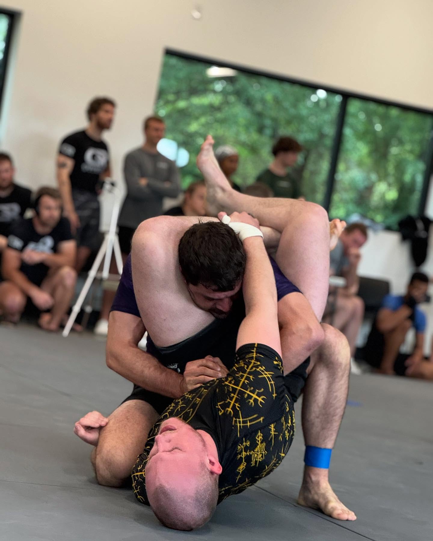 Two people grappling on the floor during a martial arts match, with spectators watching in the background.