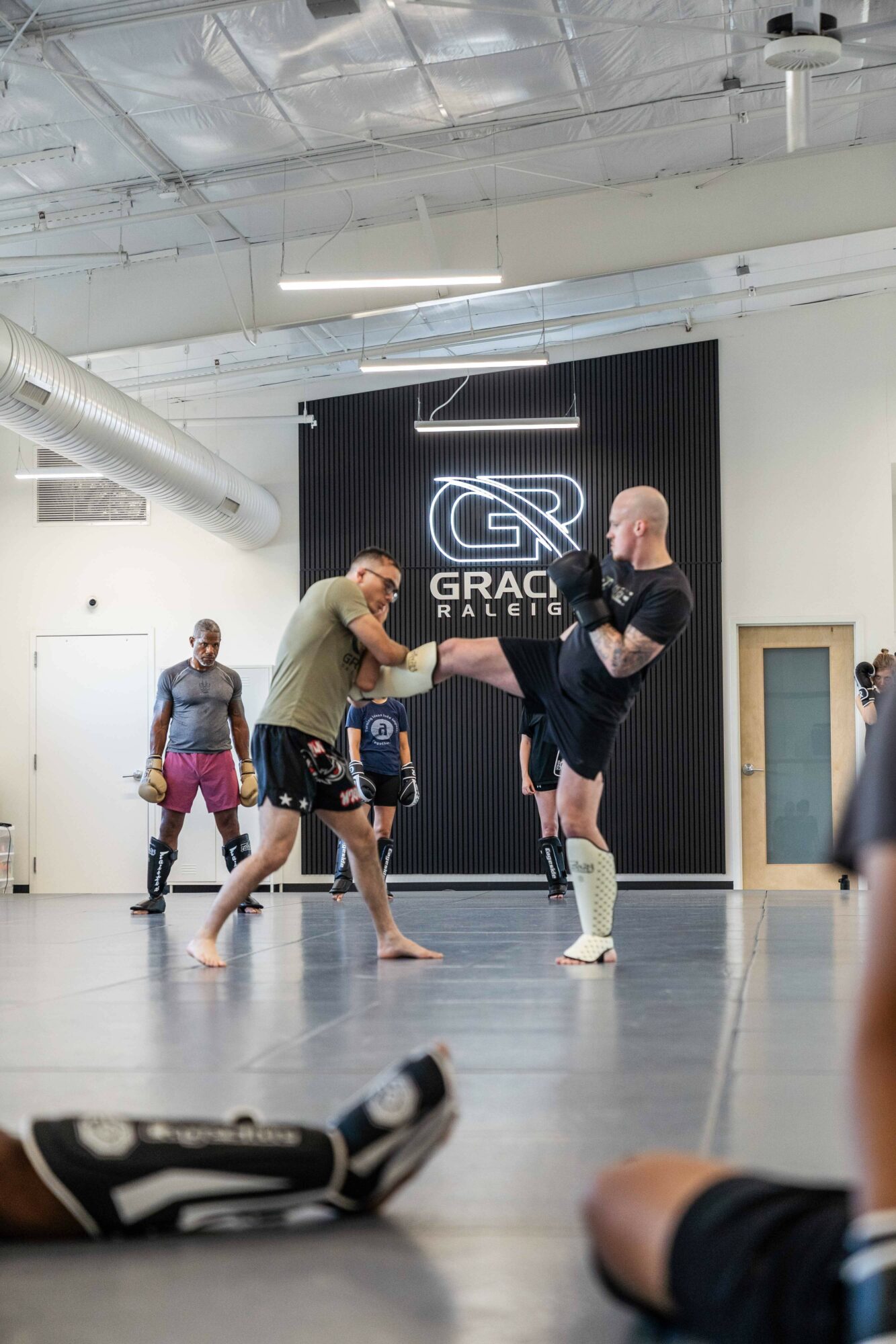 Two men practicing martial arts in a gym, one holding the other's leg, with a logo and people in the background.