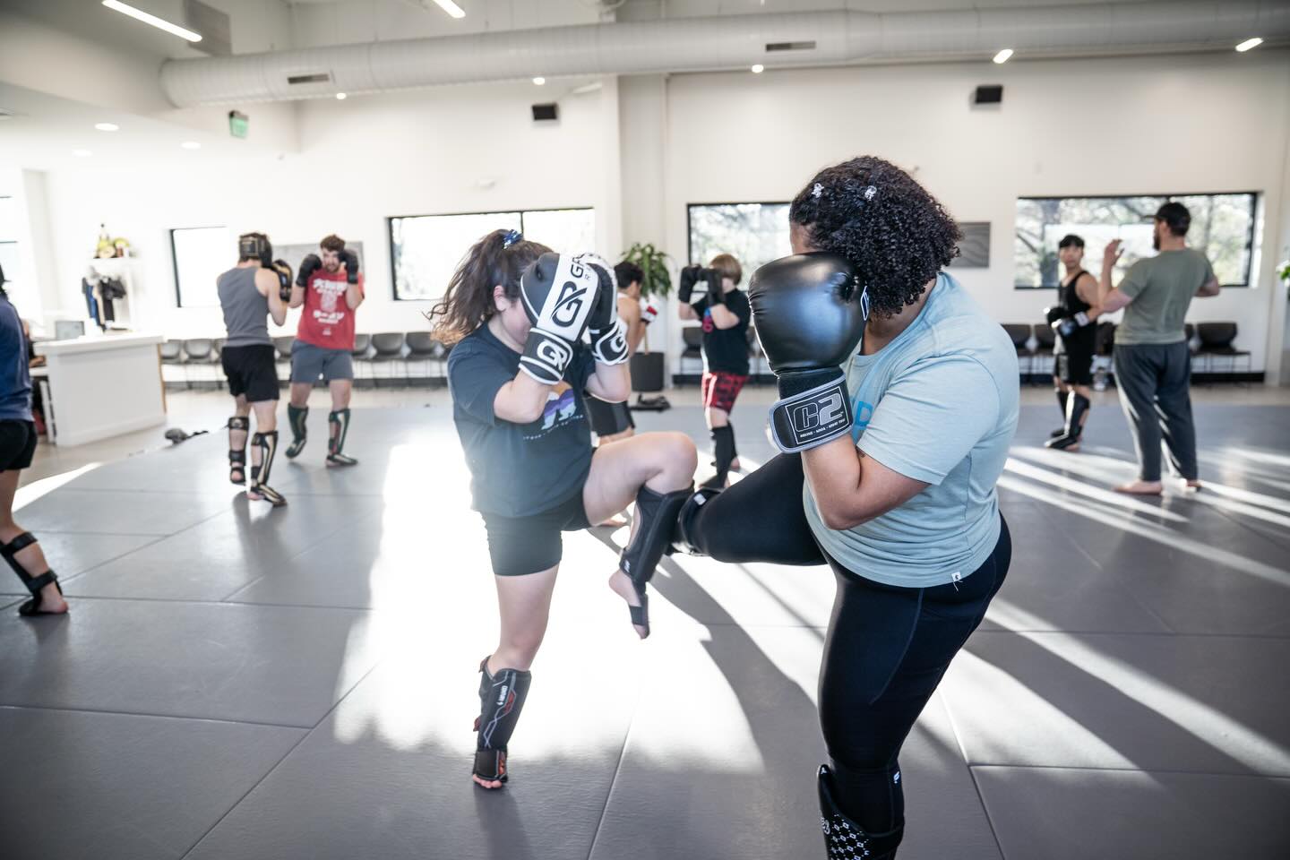 Two women sparring in a boxing gym, one wearing boxing gloves and the other kicking, with people training in the background.