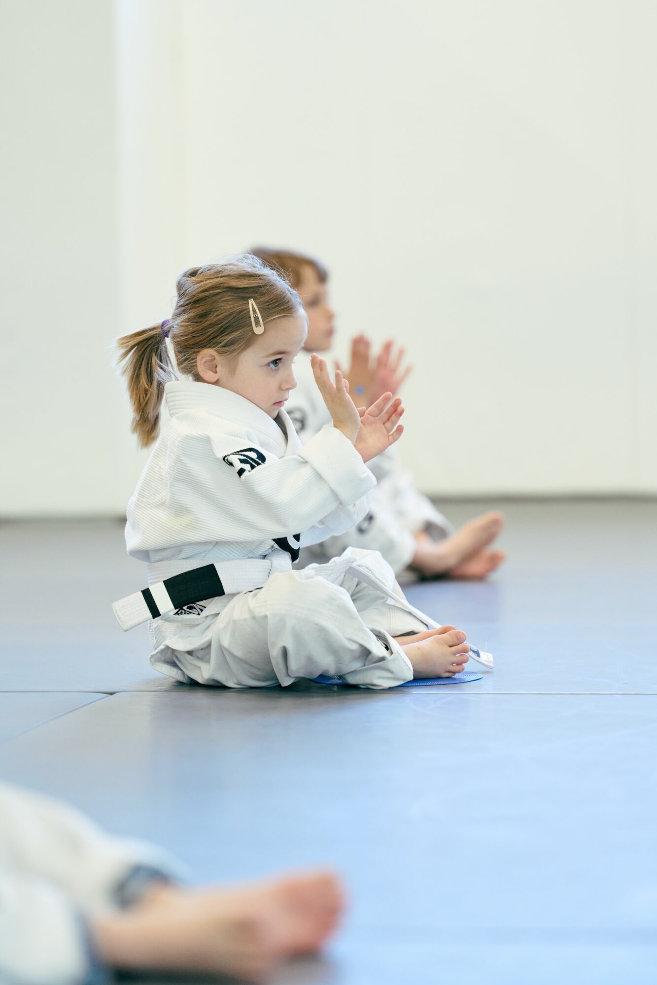 Two children in martial arts uniforms sitting on the floor, practicing techniques, with a plain white background.