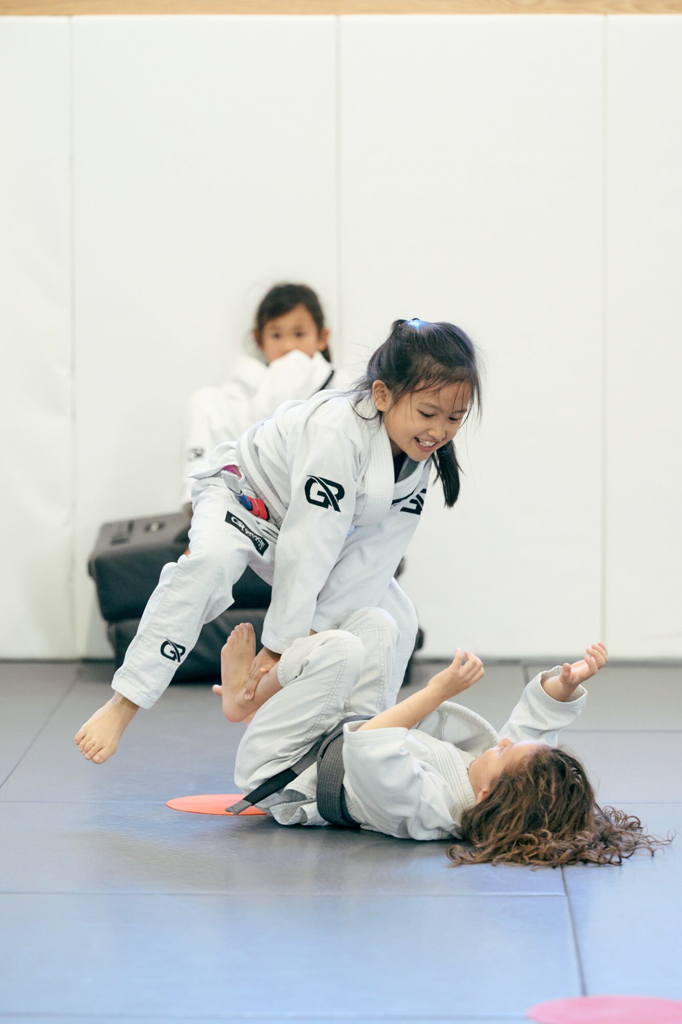 Two children practicing Brazilian Jiu-Jitsu on a blue mat, one on top of the other, with a plain white wall background.