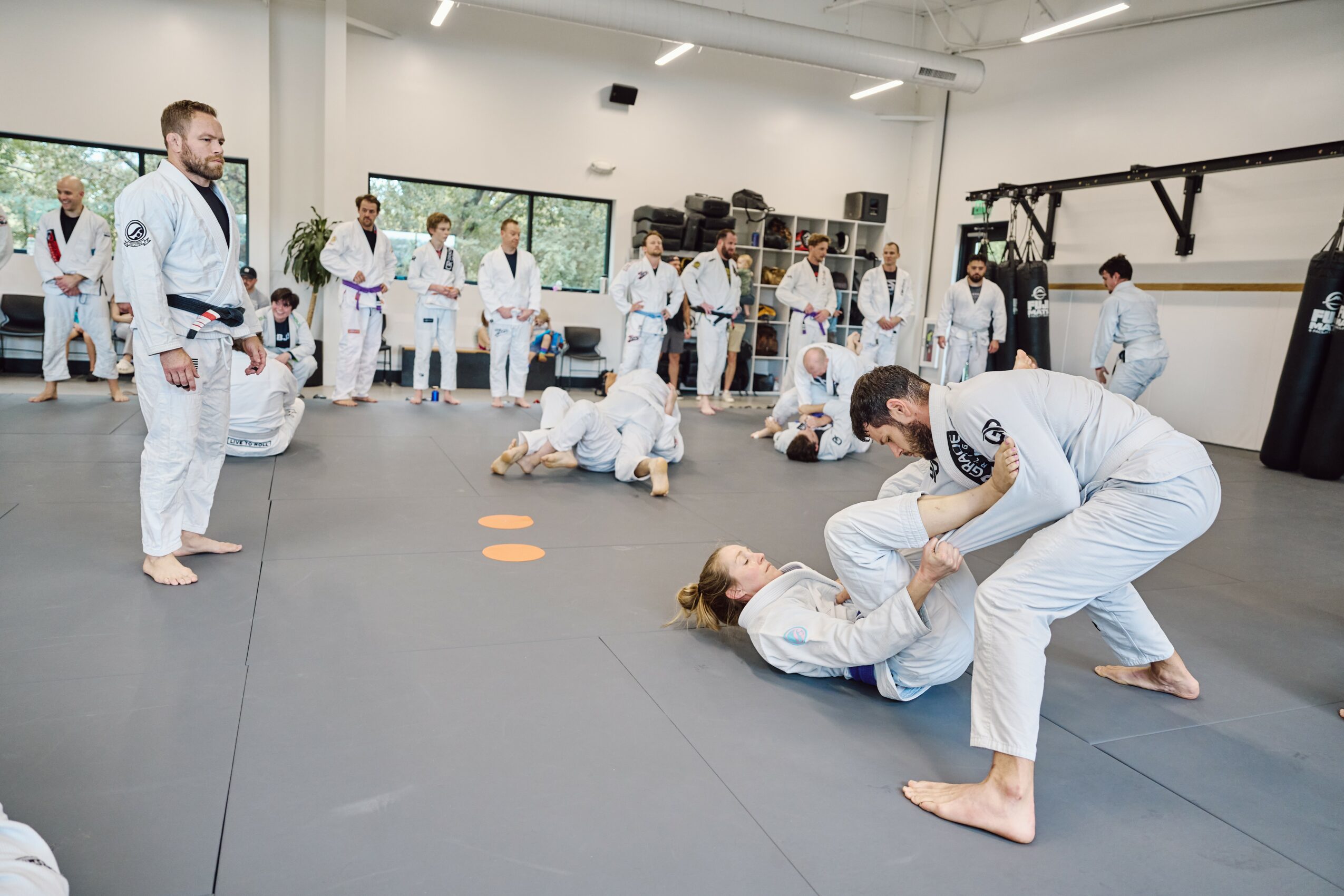 People practicing Brazilian Jiu-Jitsu in a gym, some on the ground, others observing, with a person standing nearby. Bright room with windows.