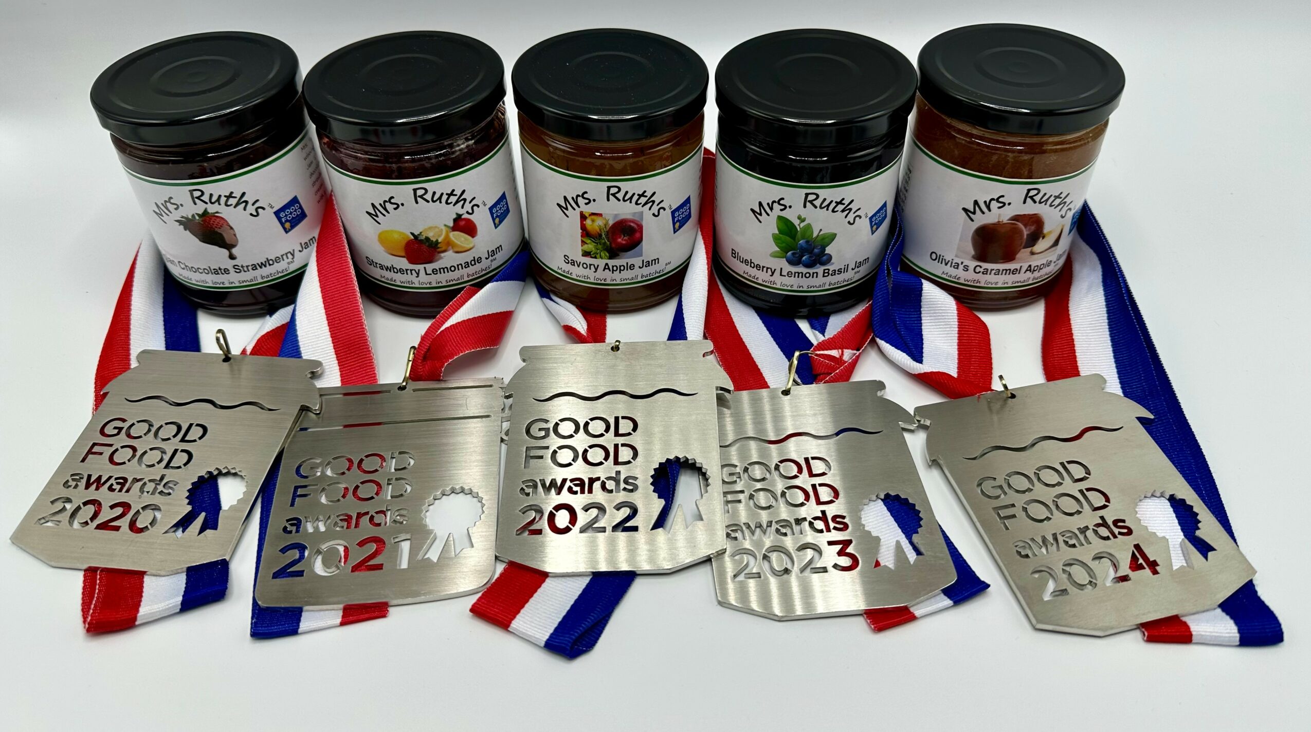 Five jars with black lids and medals with ribbons, labeled 'Good Food Awards 2022' and 2023, arranged in a row.