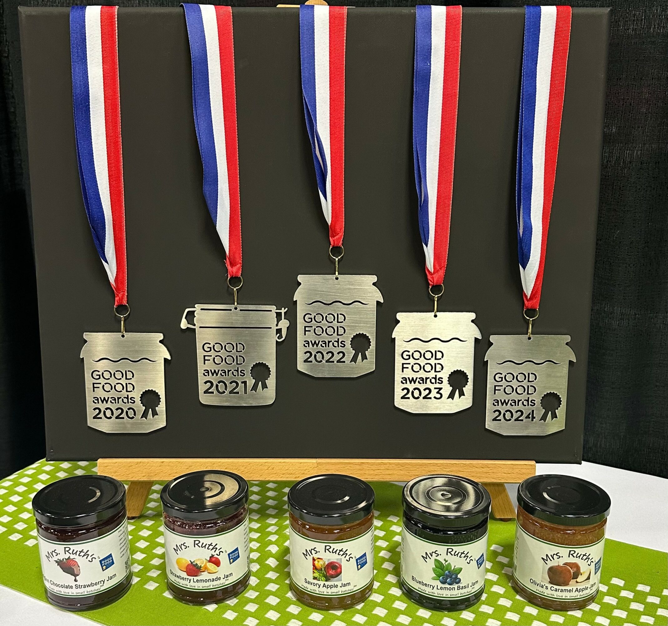 Medals with ribbons and jars of food products on a green surface, awards from 2020 to 2024, with a black background.