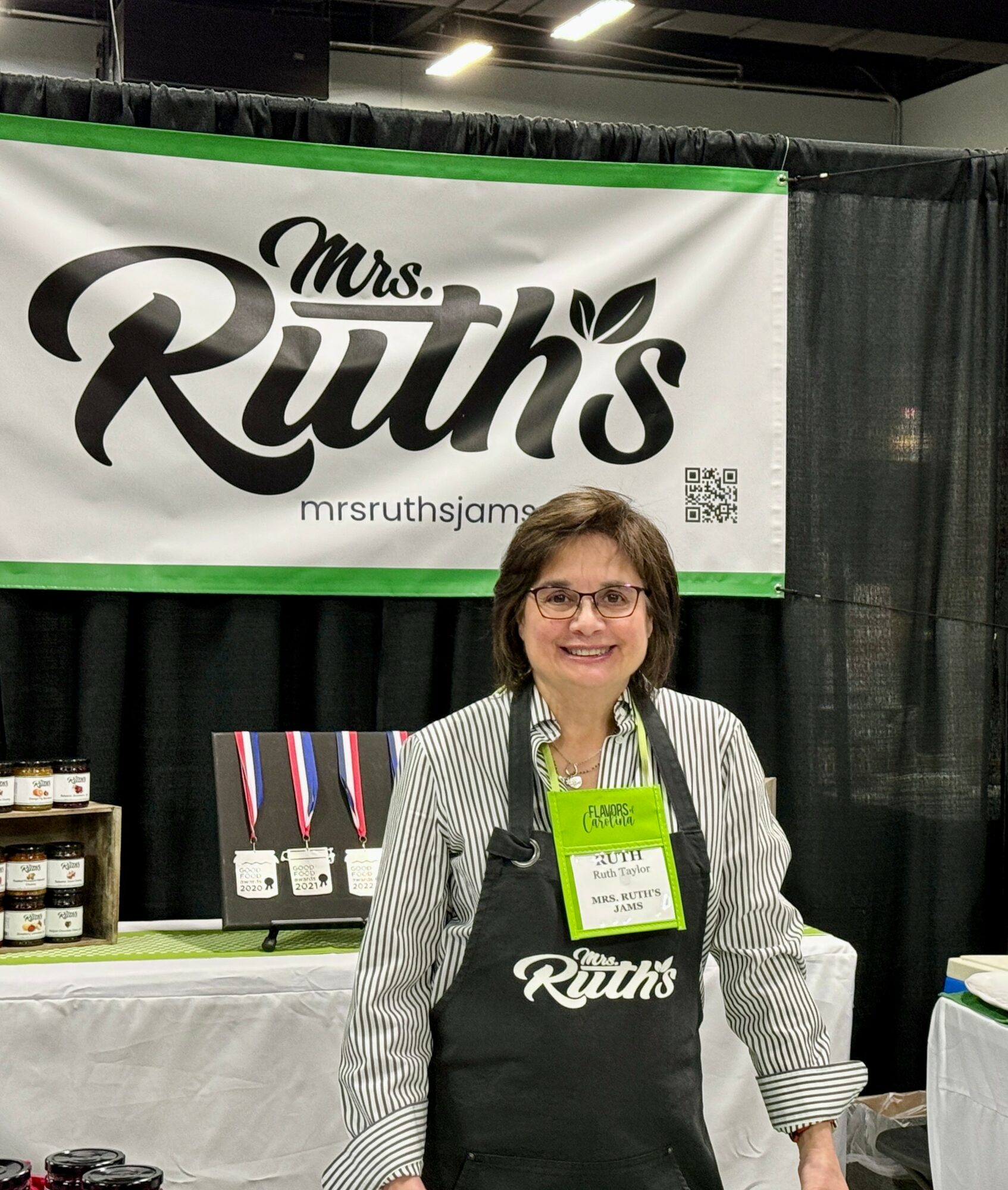 Smiling woman wearing glasses and an apron stands in front of a booth with a Mrs. Ruth's banner at an indoor event.