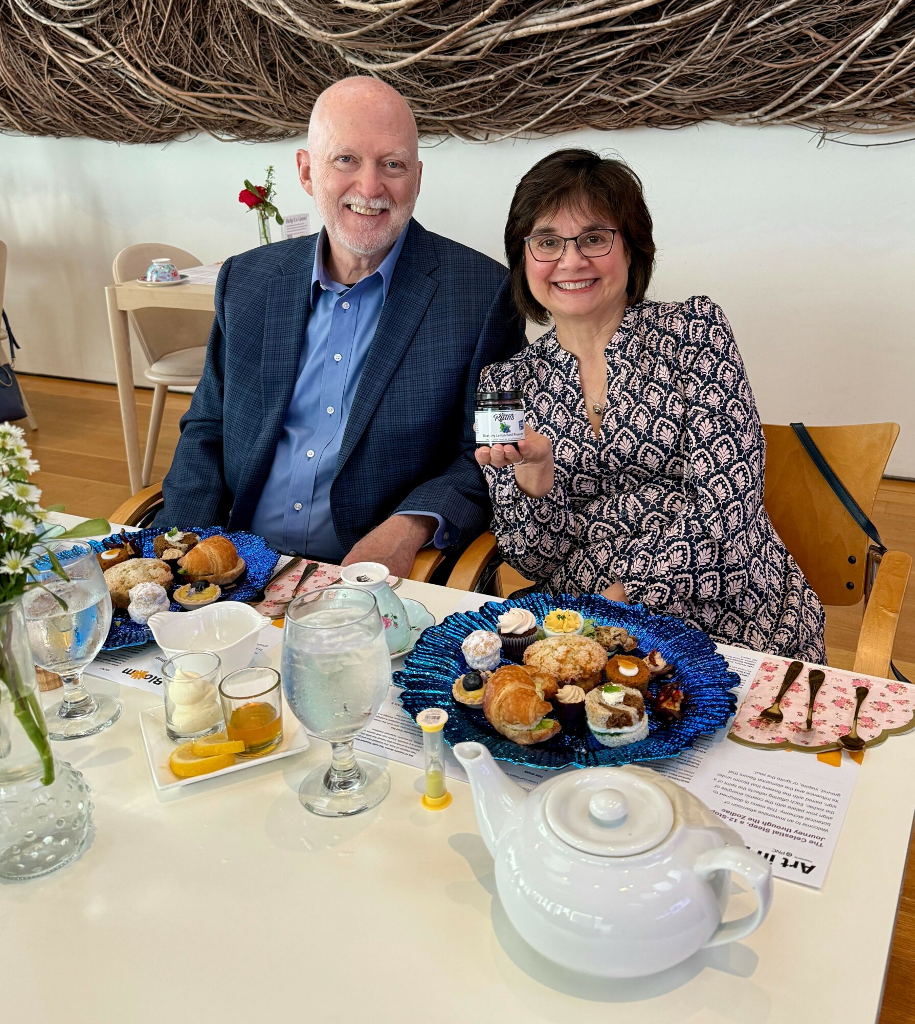 Two people sitting at a table with desserts and drinks, smiling, in a bright room with a white wall and wooden furniture.