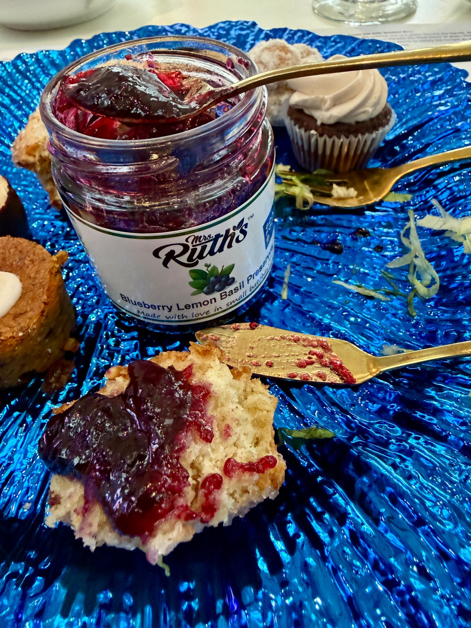 Jar of berry jam, cupcake, and a piece of cake on a blue plate with gold utensils, some crumbs visible.