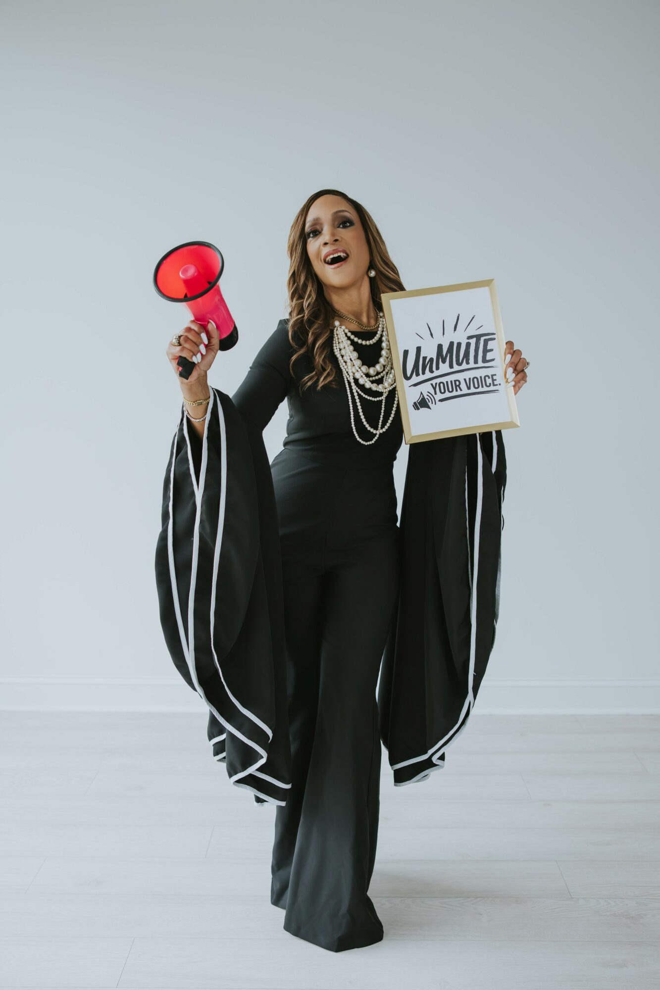 Woman in black suit holding a red megaphone and a sign that says 'UnMute Your Voice' with white background.