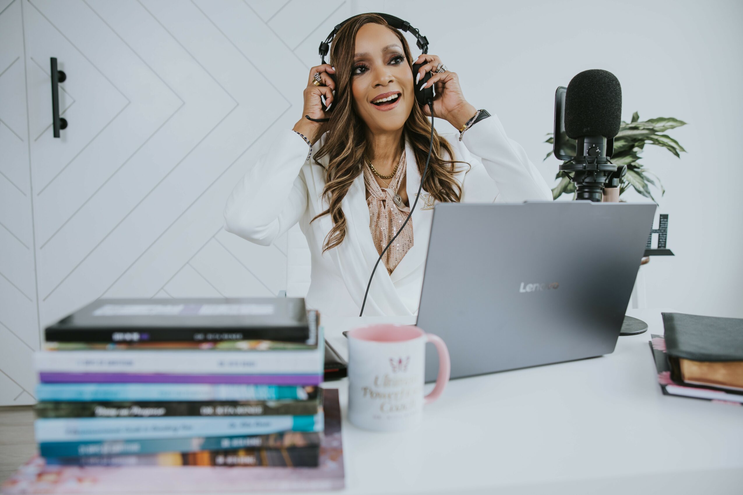 Woman with headphones smiling at laptop in a bright workspace, with books, a mug, and a plant on the desk.