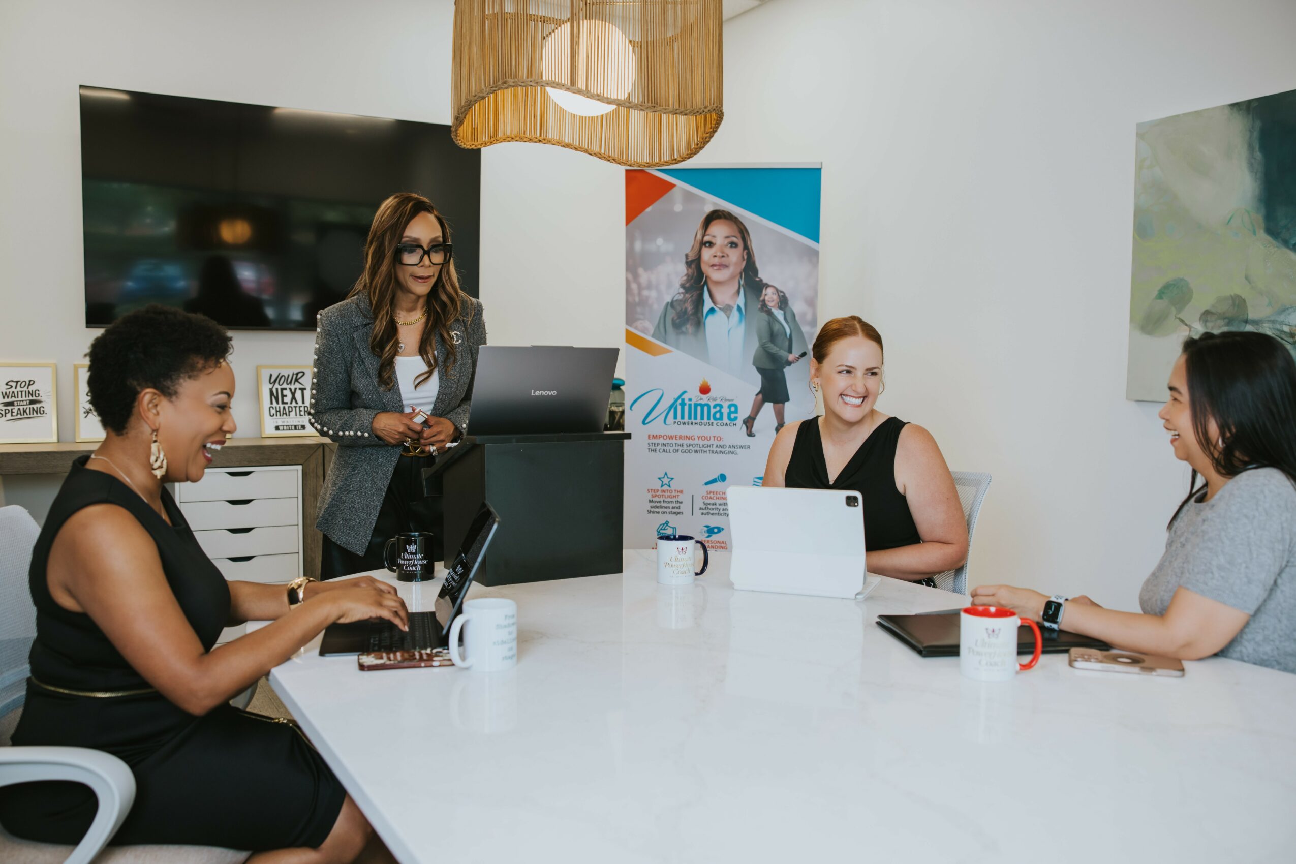 Four women seated around a white table in a meeting room, with a woman standing at a counter, using a laptop.
