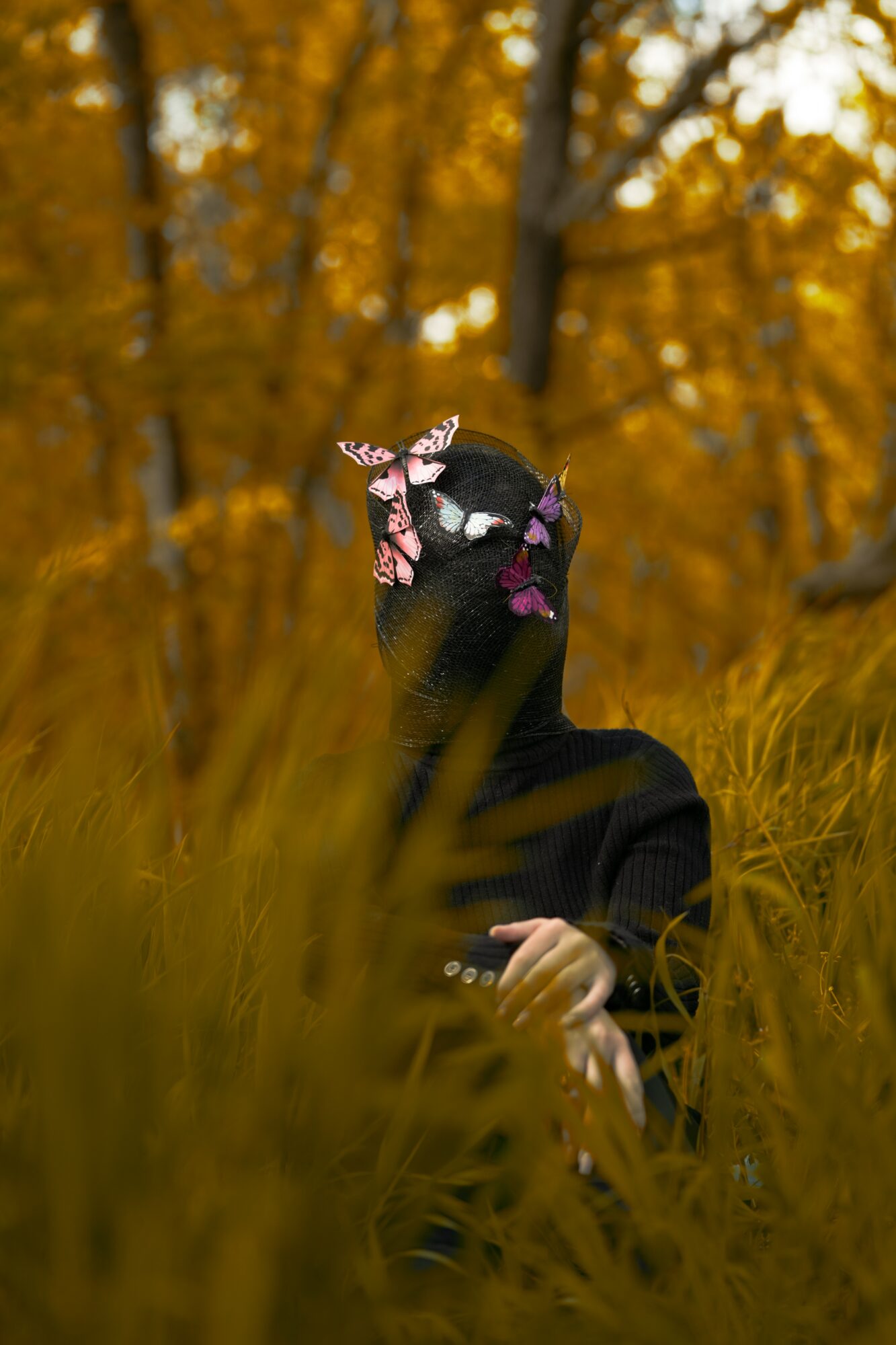 Person sitting in tall grass surrounded by autumn trees, wearing a black outfit and a colorful headscarf.