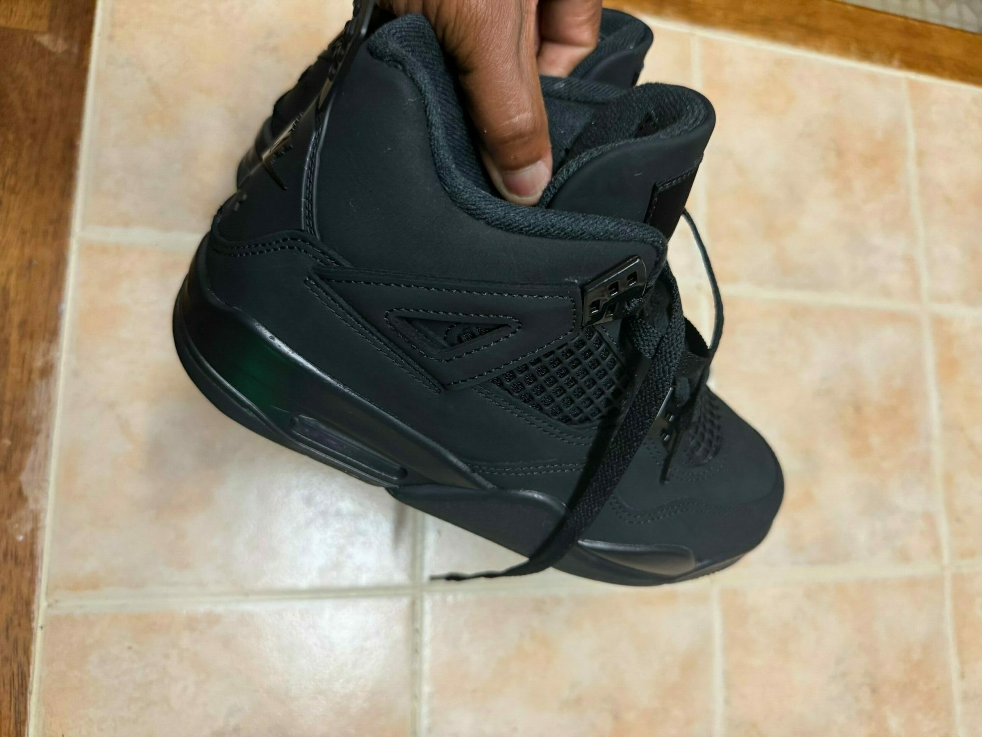 Person holding a black athletic shoe against a tiled wall, with the shoe's sole facing right.