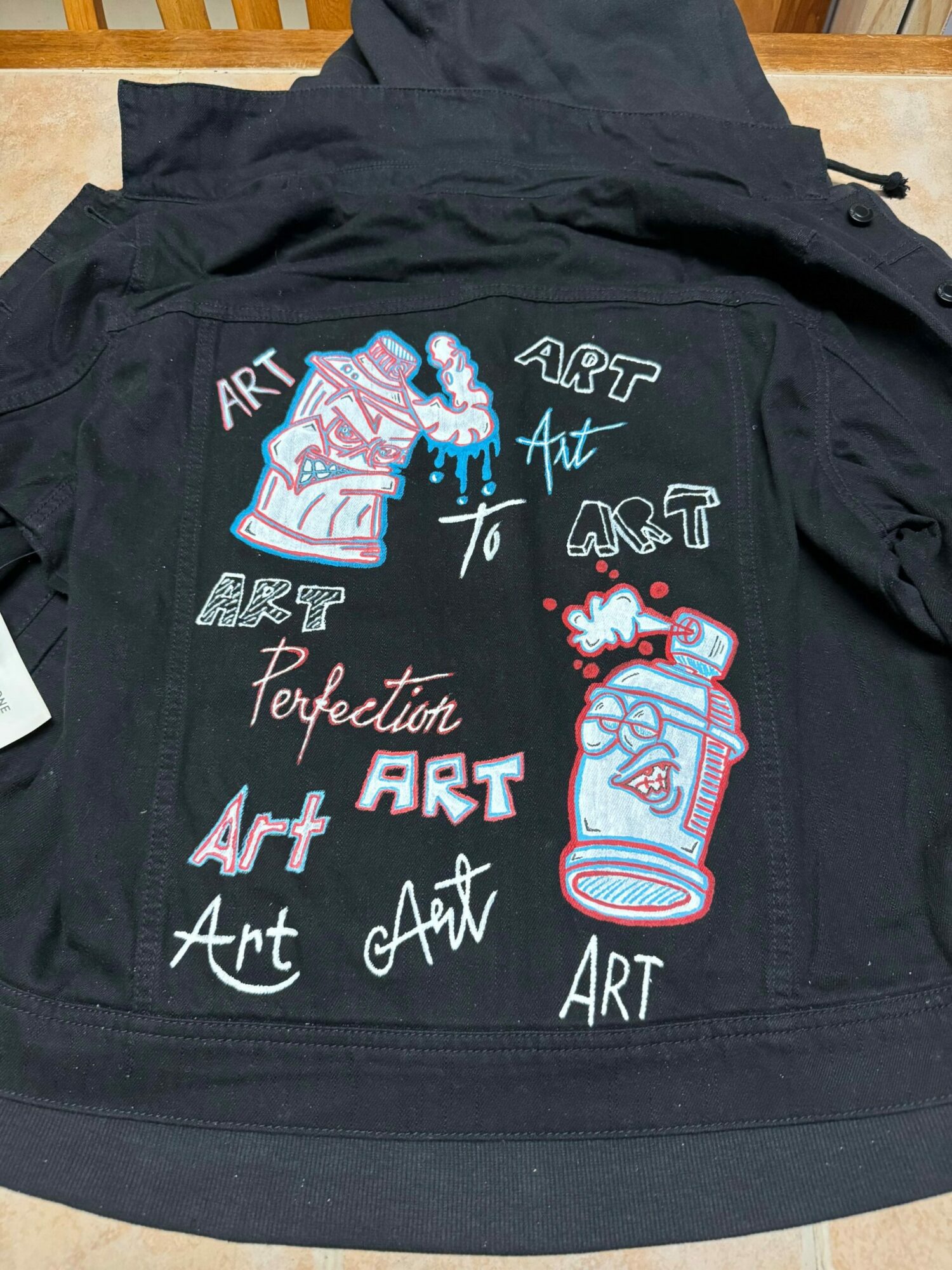 Black backpack with colorful embroidered art designs and text, including a paint can, paintbrush, and phrases about art, on a wooden surface.