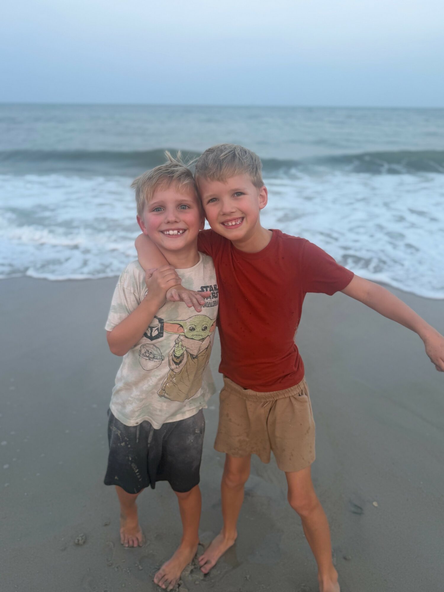 Two children smiling and hugging on the beach with ocean waves in the background.
