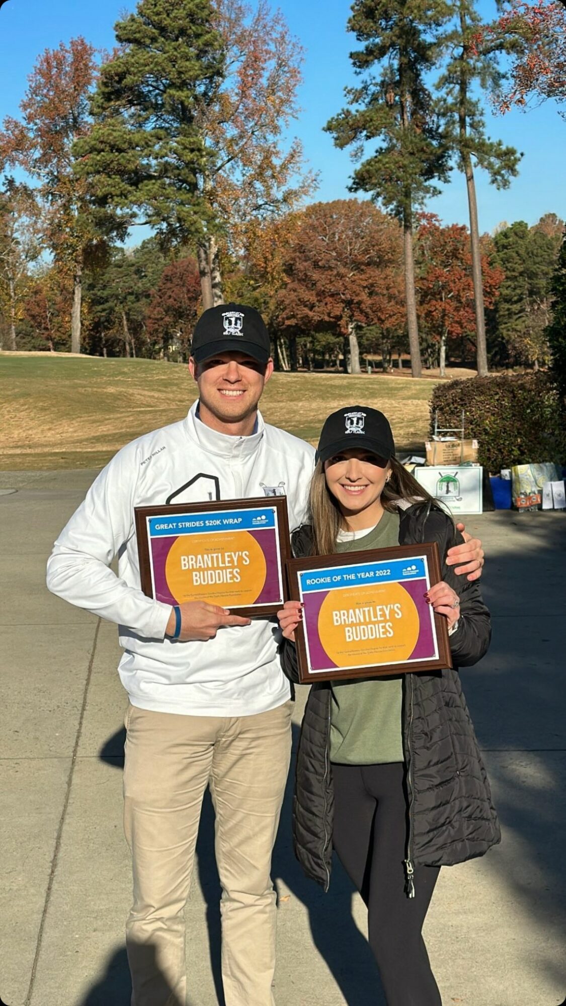 Two people outdoors holding certificates, smiling, with trees and a park in the background.