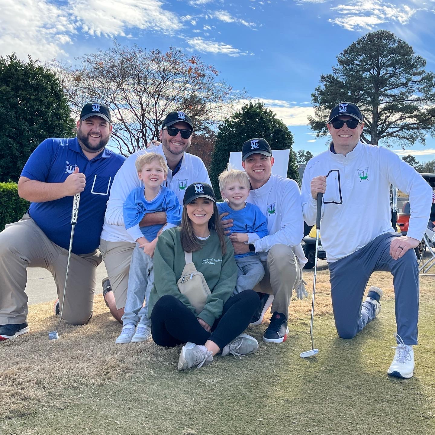 Group of six people, including children, posing outdoors on a golf course with trees and blue sky in background.