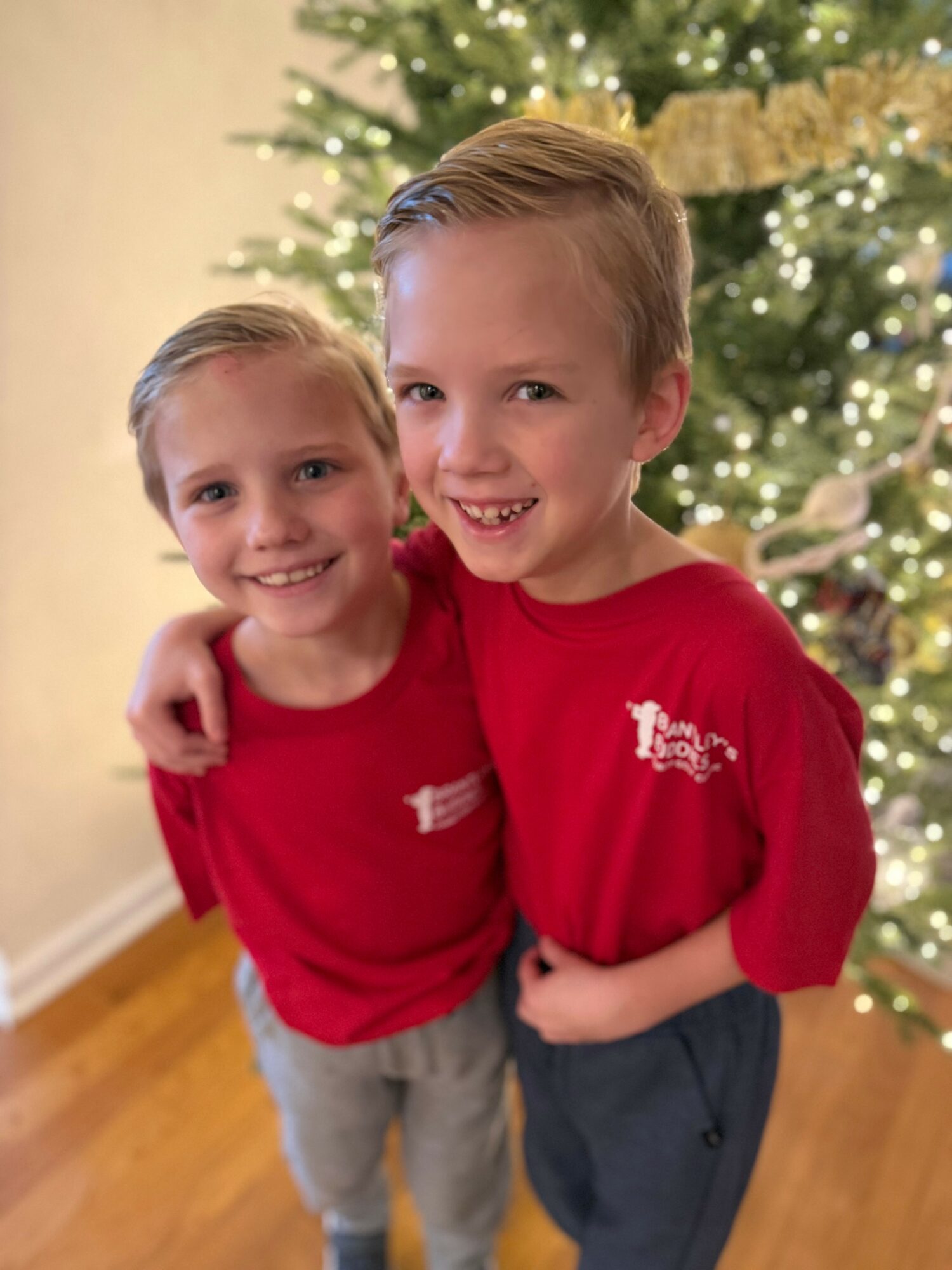 Two children smiling, standing close, in front of a decorated Christmas tree, wearing red shirts, indoors.