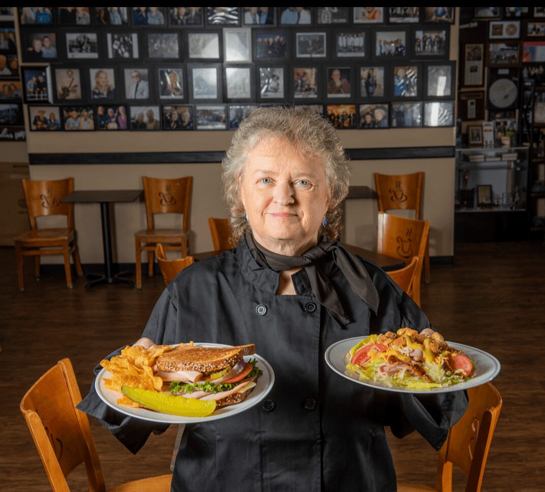 Older woman in black chef uniform holding two plates of food, standing in a restaurant with wooden chairs and photo collage on wall.