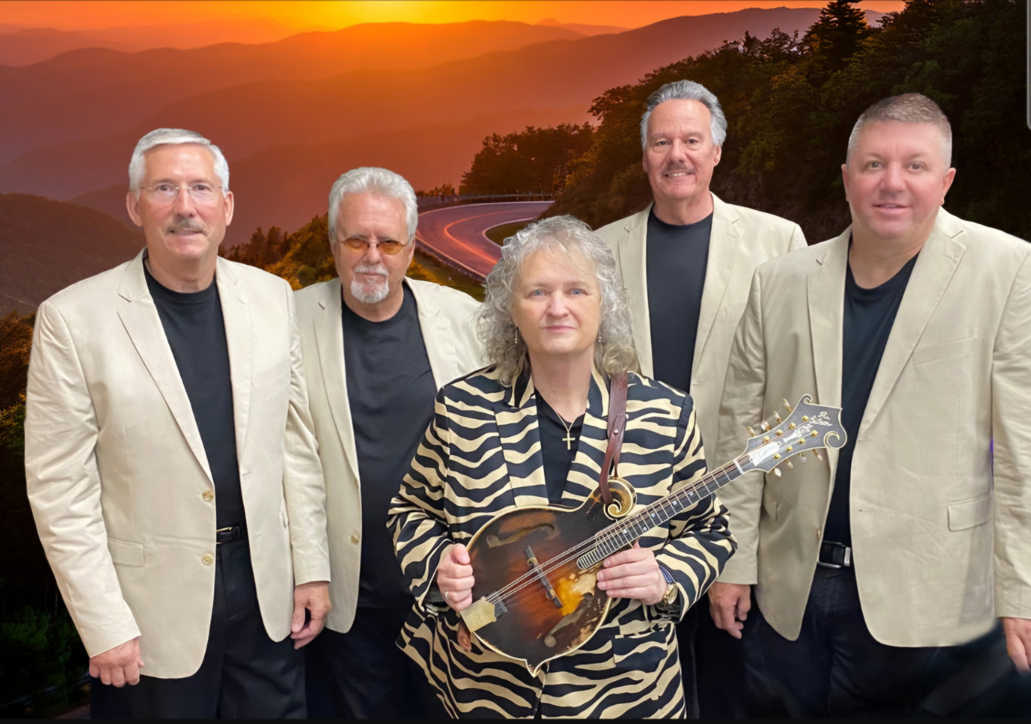 Group of five people, four men and one woman, standing outdoors with a sunset and mountain landscape in the background. The woman holds a guitar.