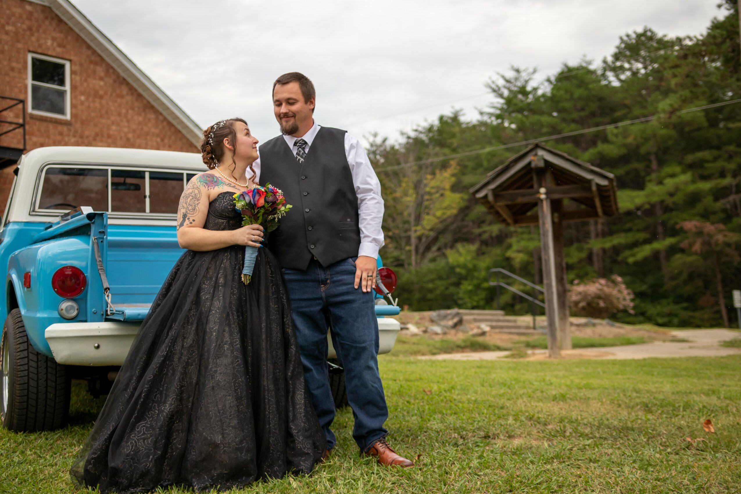 A couple dressed in formal attire standing outdoors near a blue vintage truck, smiling and holding a bouquet of flowers.
