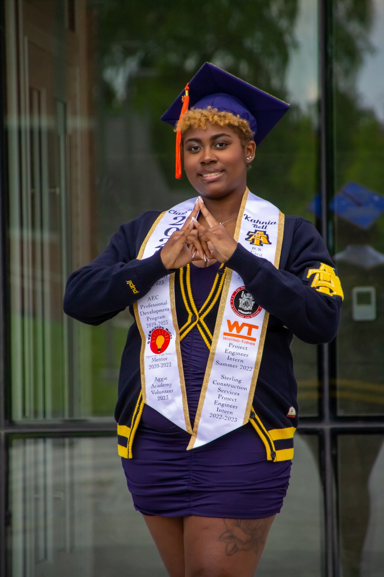 Graduate in cap and gown with multiple sashes, standing outdoors with hands clasped, smiling.