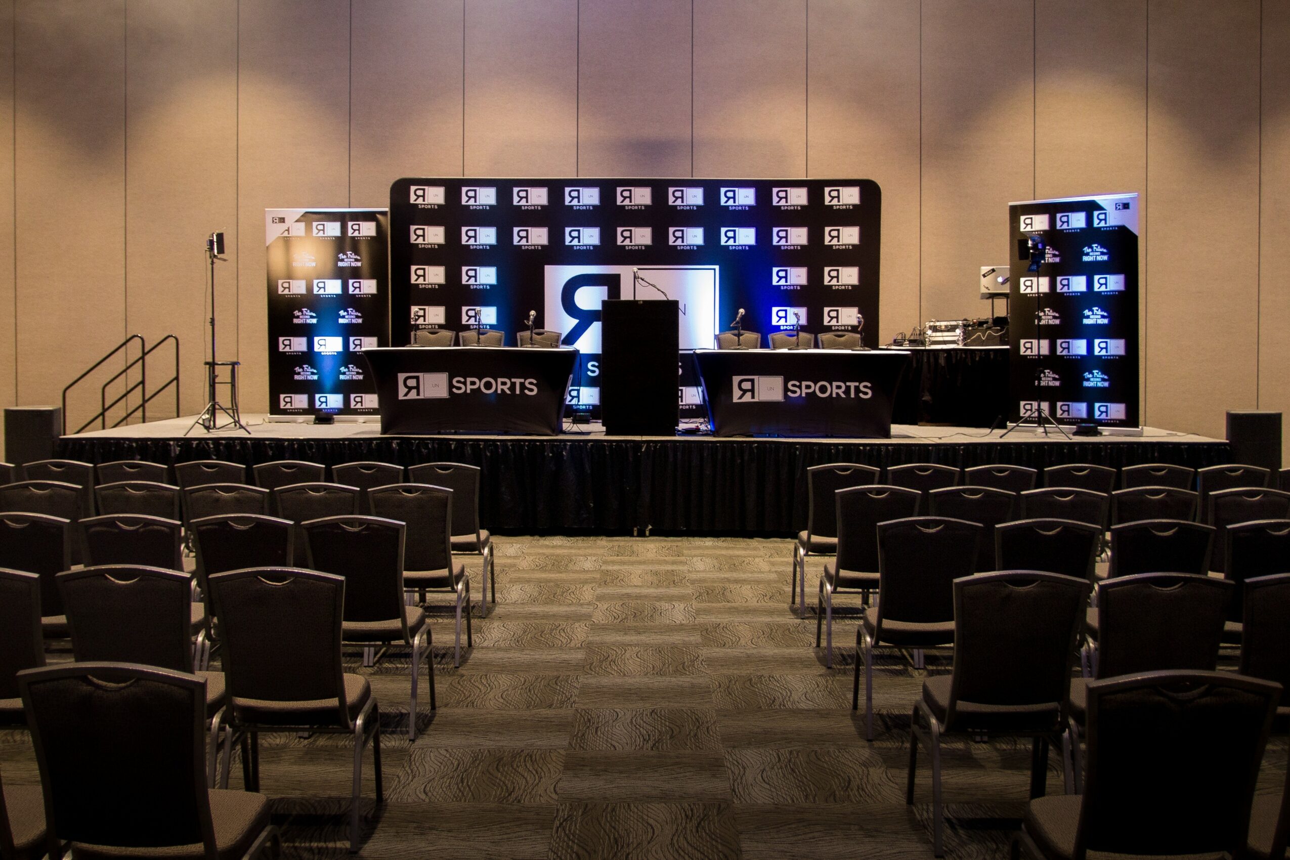 Empty conference room with chairs facing a stage with screens and banners, set up for a sports event or presentation.