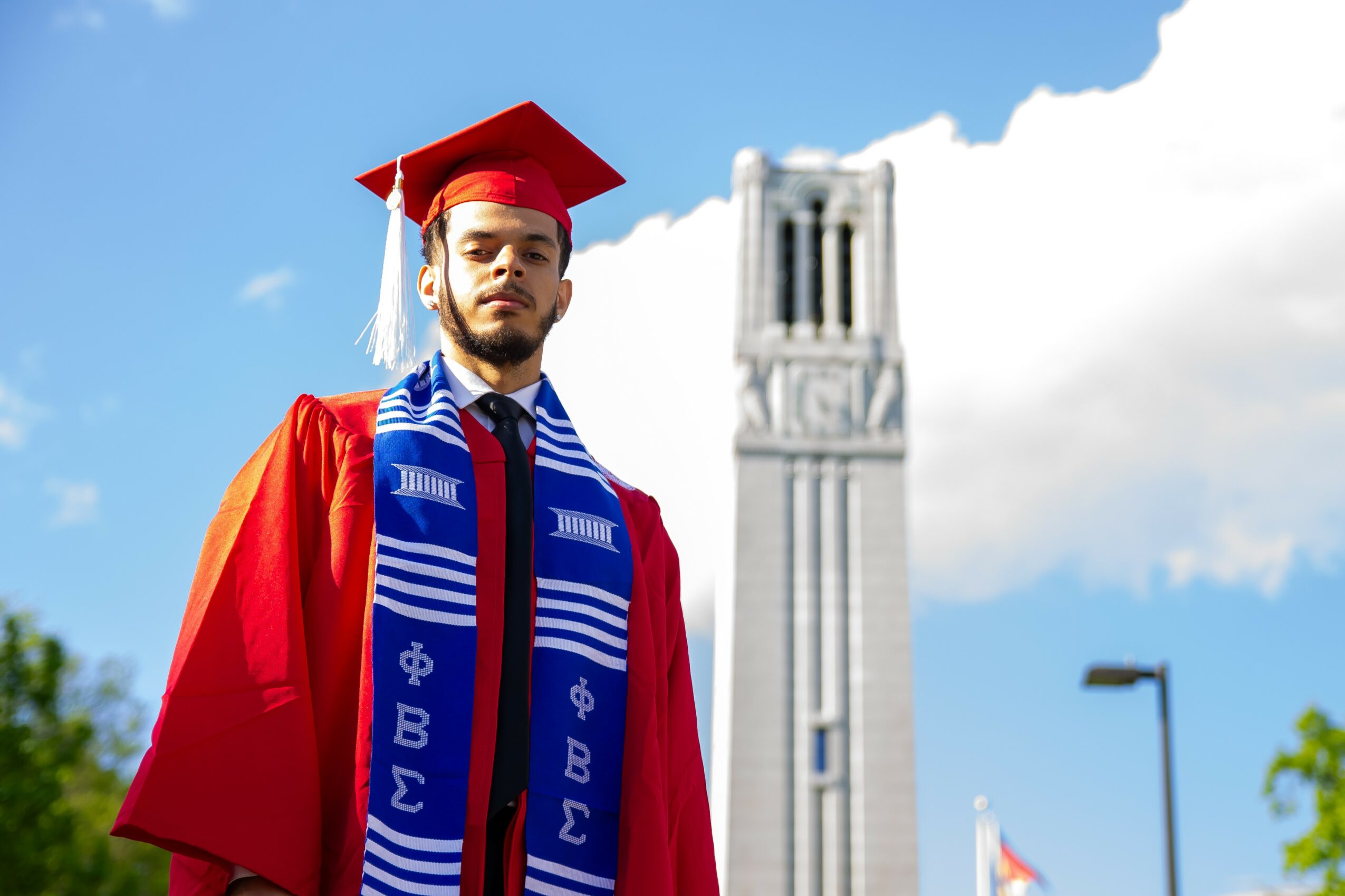 Graduate in cap and gown standing outdoors with a tall clock tower in the background, blue sky, and clouds.