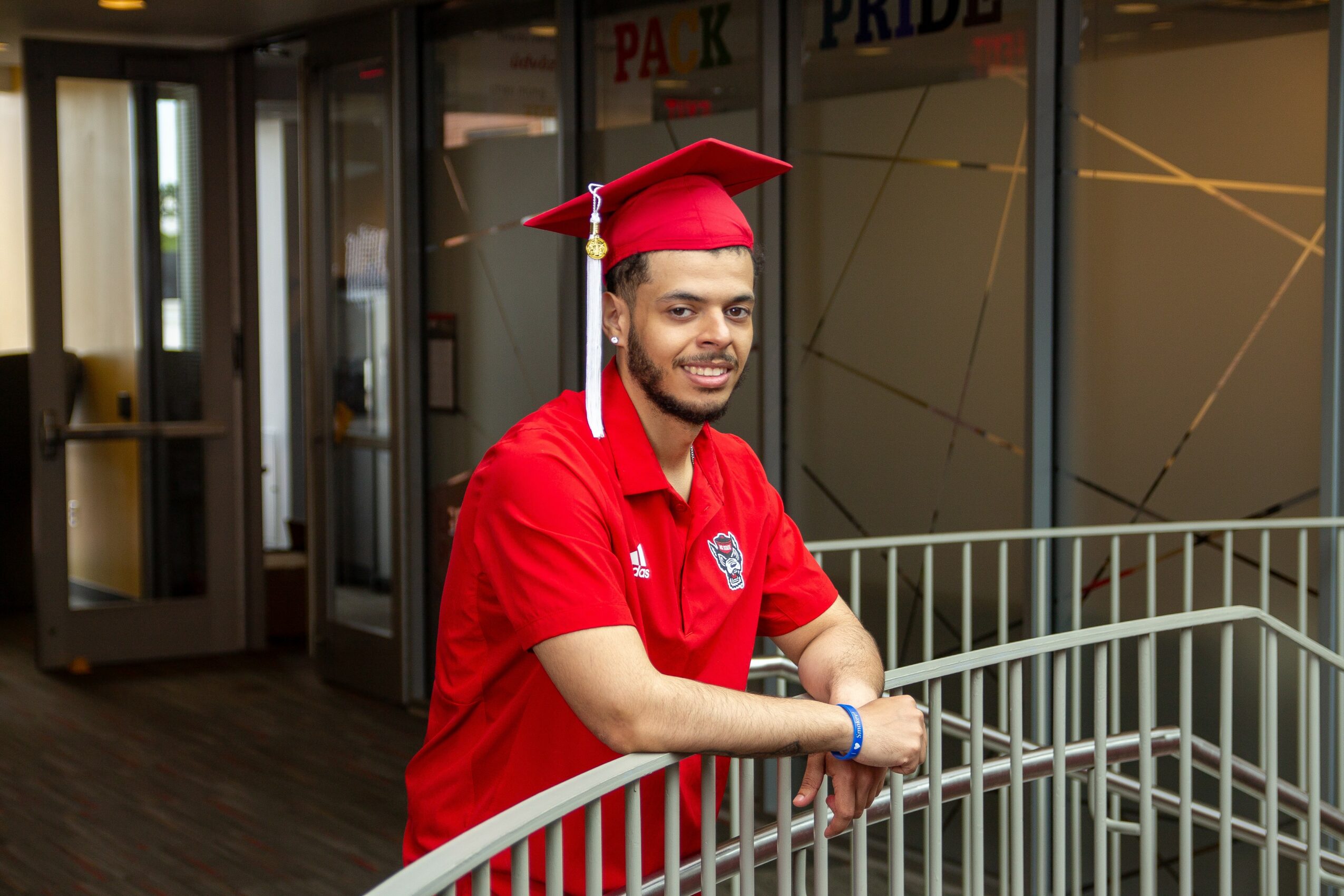 Young man in red graduation cap and shirt leaning on a metal railing indoors, smiling, with glass doors behind him.