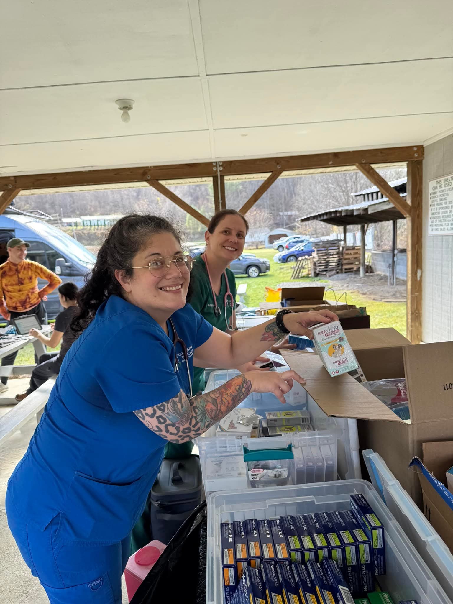 Two women smiling and handing out items at an outdoor event, with tables and boxes in front of them.