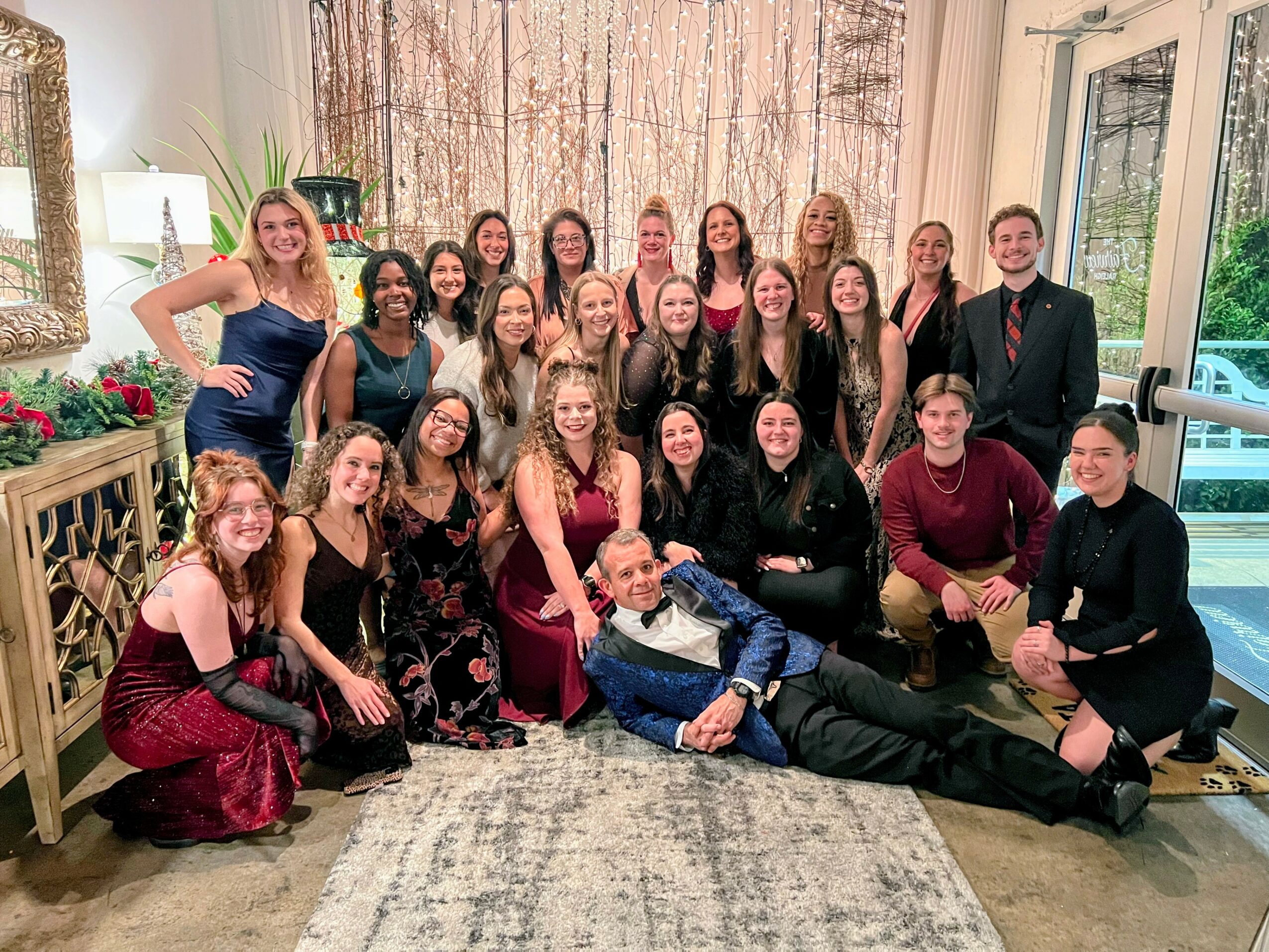 Group of people posing indoors, some standing and some sitting, with a window and curtains in the background.
