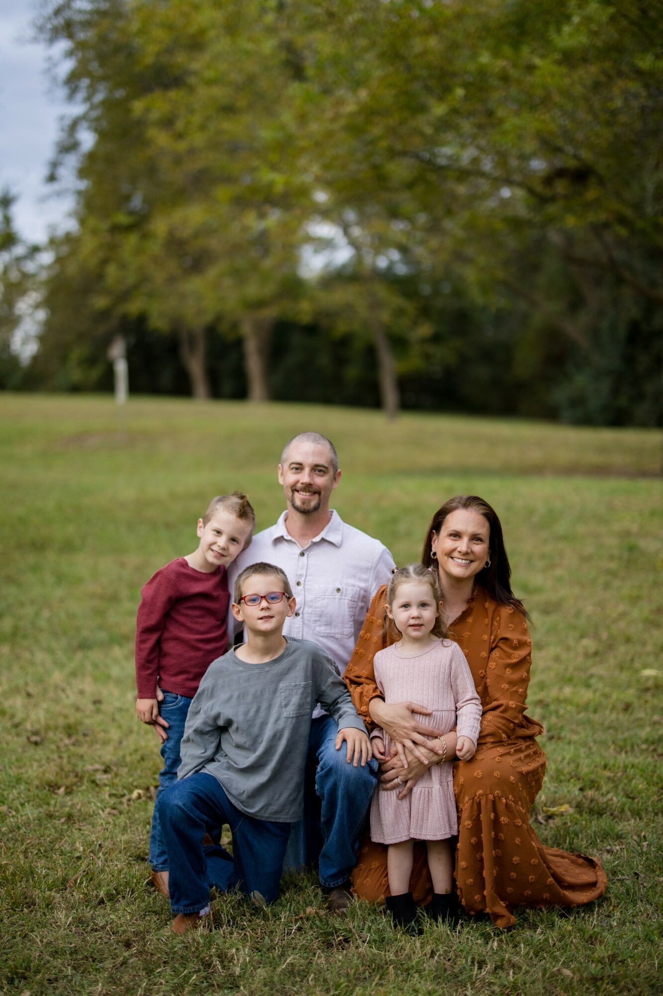 Family of five outdoors on grass with trees in background, smiling, posing for photo.