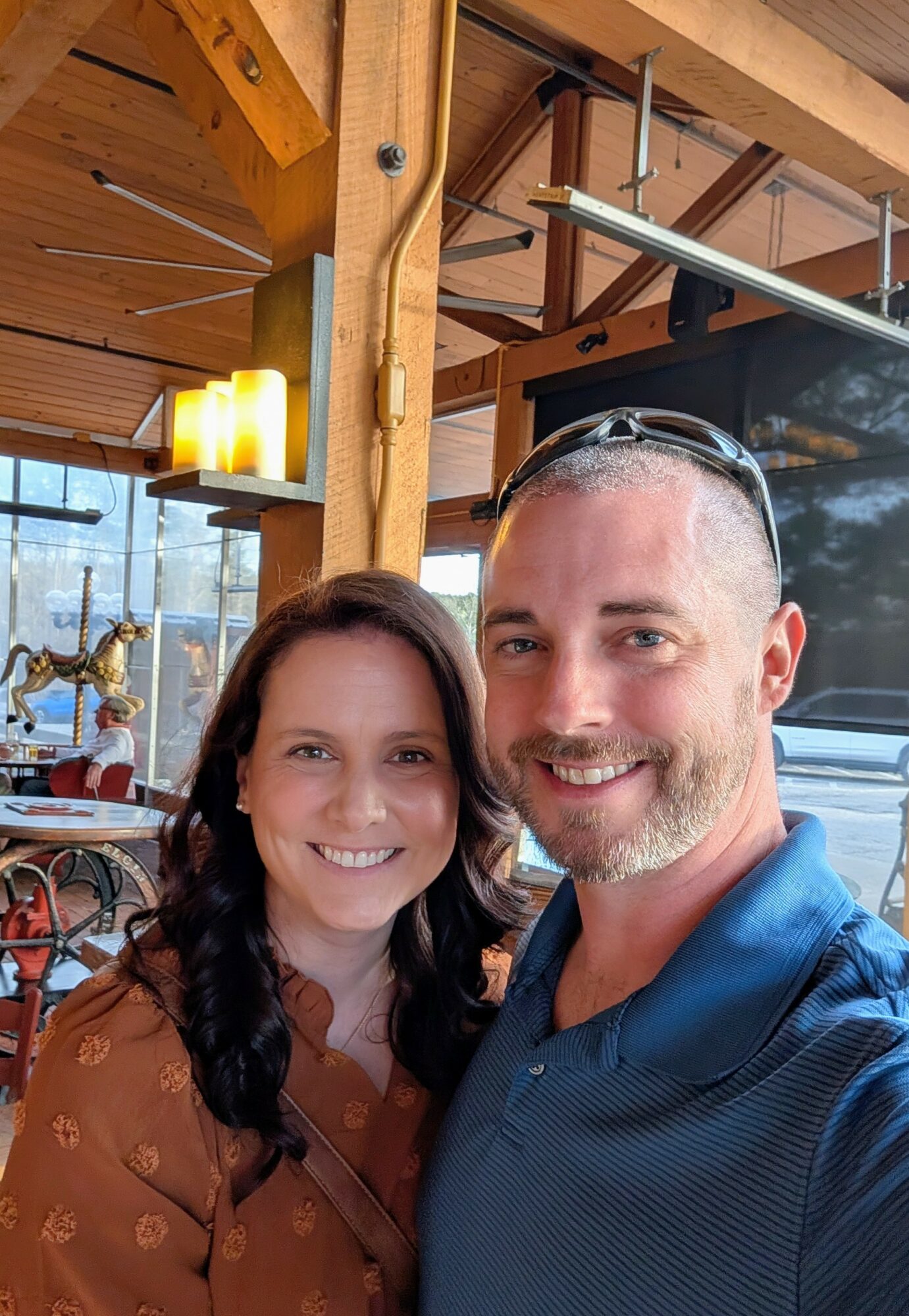 Two people smiling indoors with wooden beams and decorative items in the background.