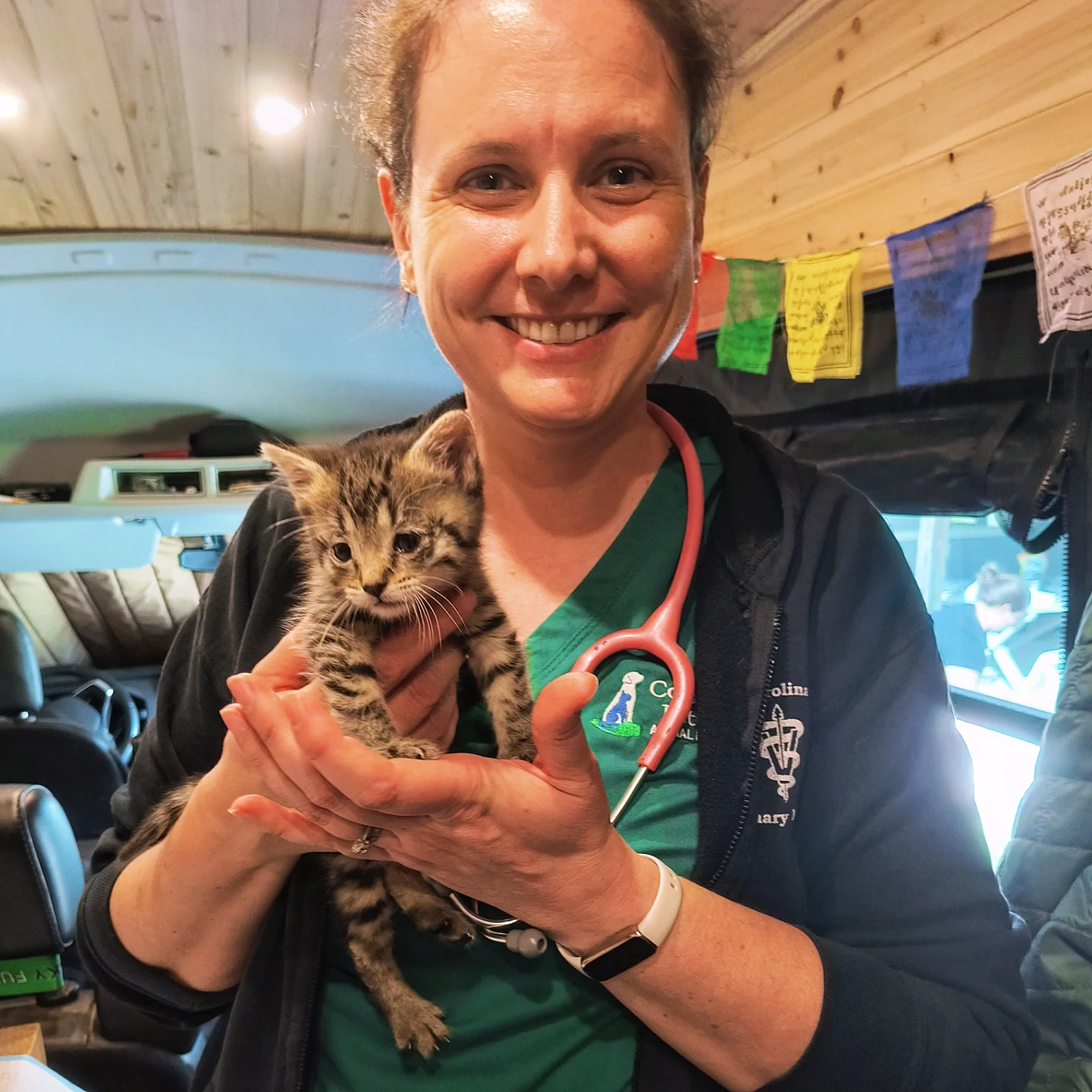 Woman holding a small tabby kitten inside a vehicle, smiling at the camera, with colorful flags hanging in the background.