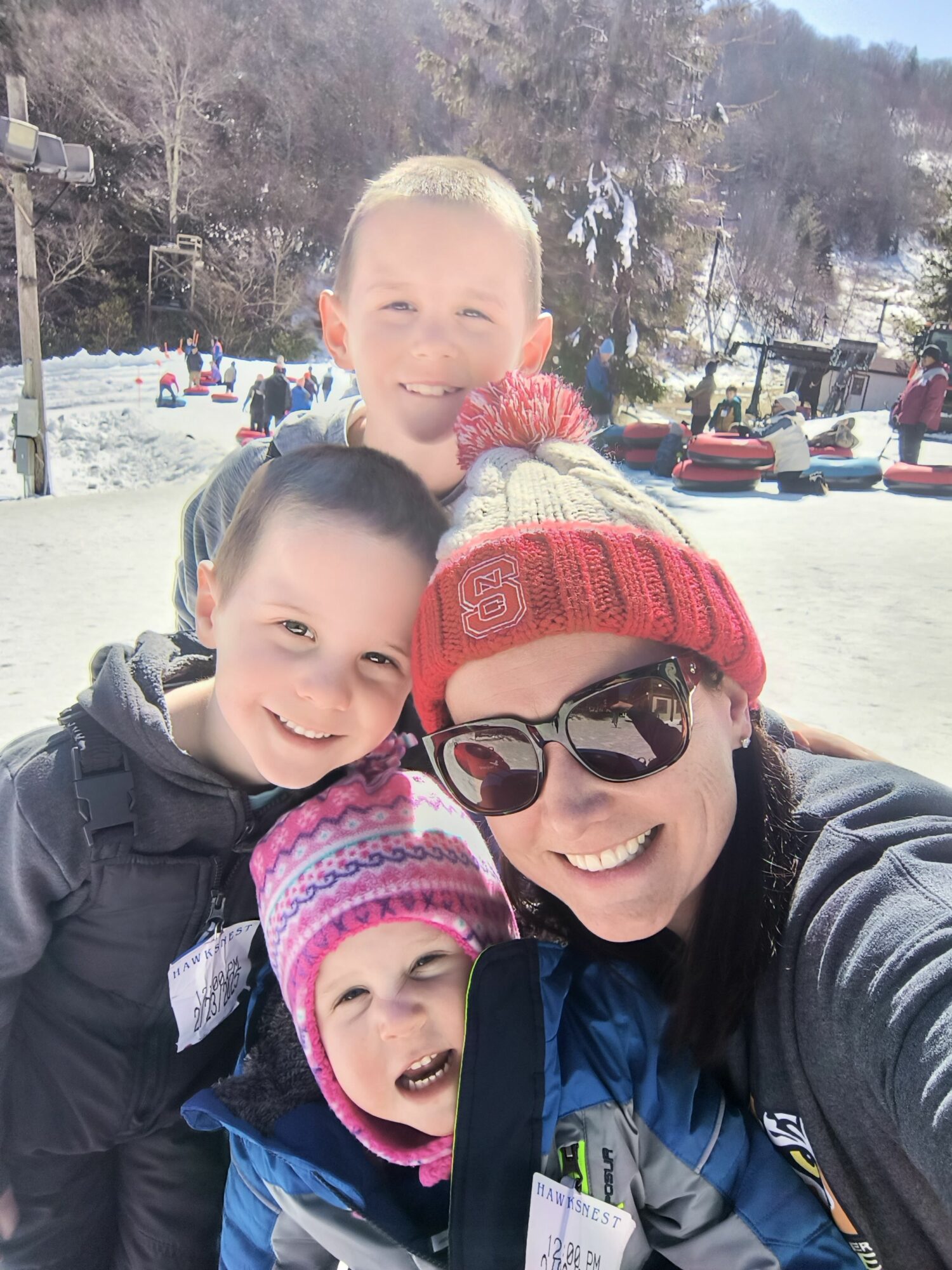 Four people outdoors, smiling, bundled in winter clothing, with snow and trees in the background.