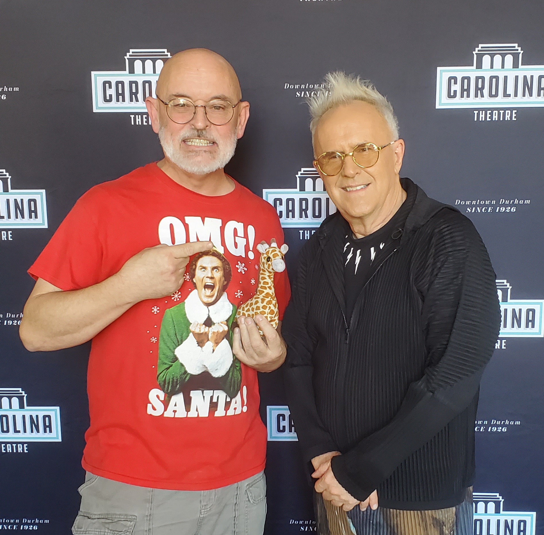 Two men standing in front of a backdrop, one wearing a red Christmas sweater and the other in a black jacket, smiling.