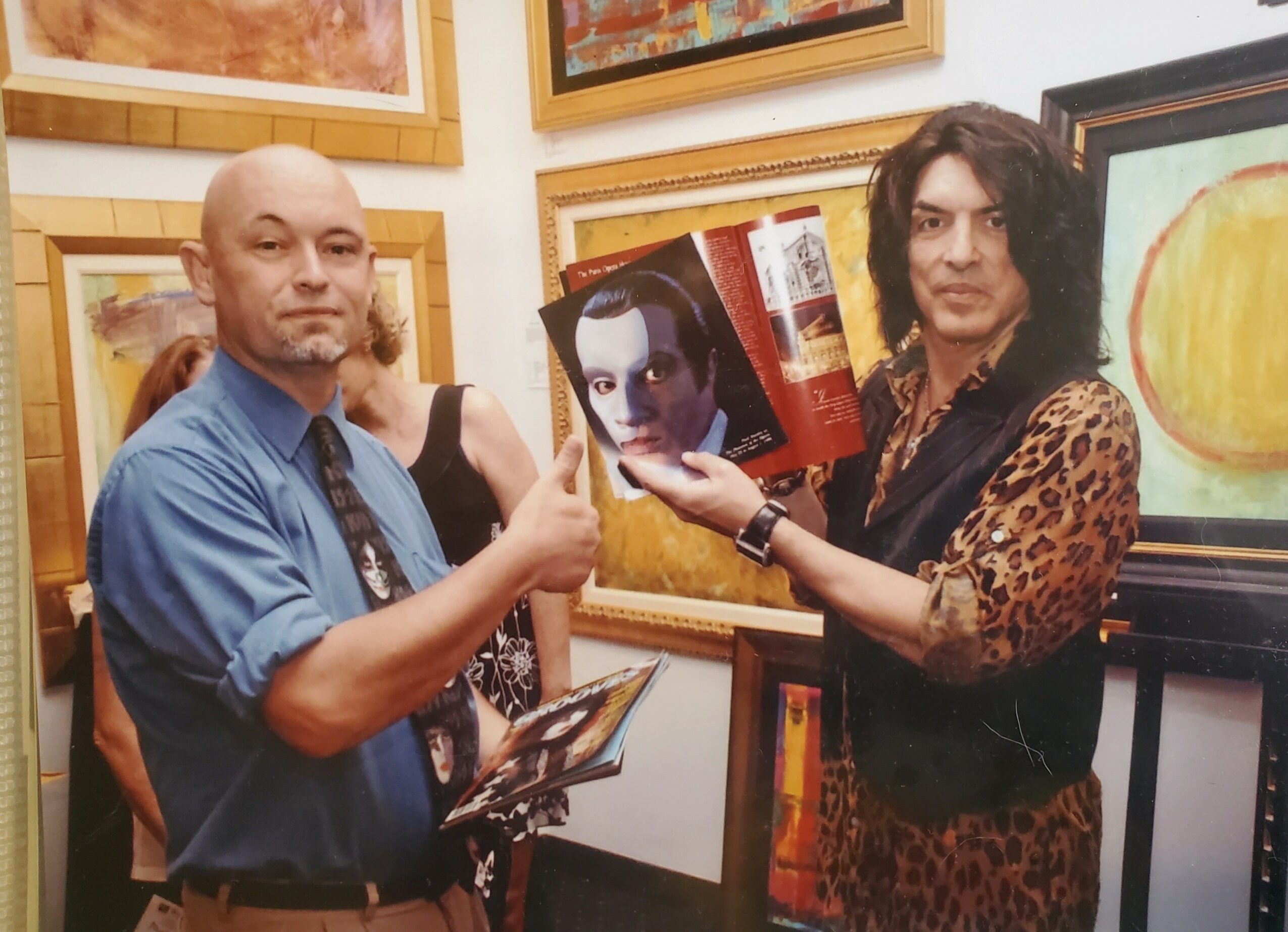 Two people at an art gallery, one holding a book with a portrait, surrounded by framed artwork.