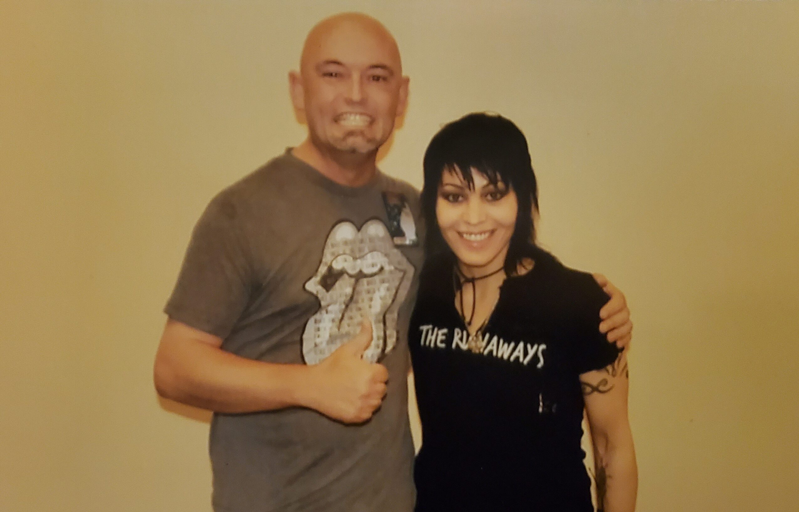 Two people standing close together, smiling, with a plain light-colored background.