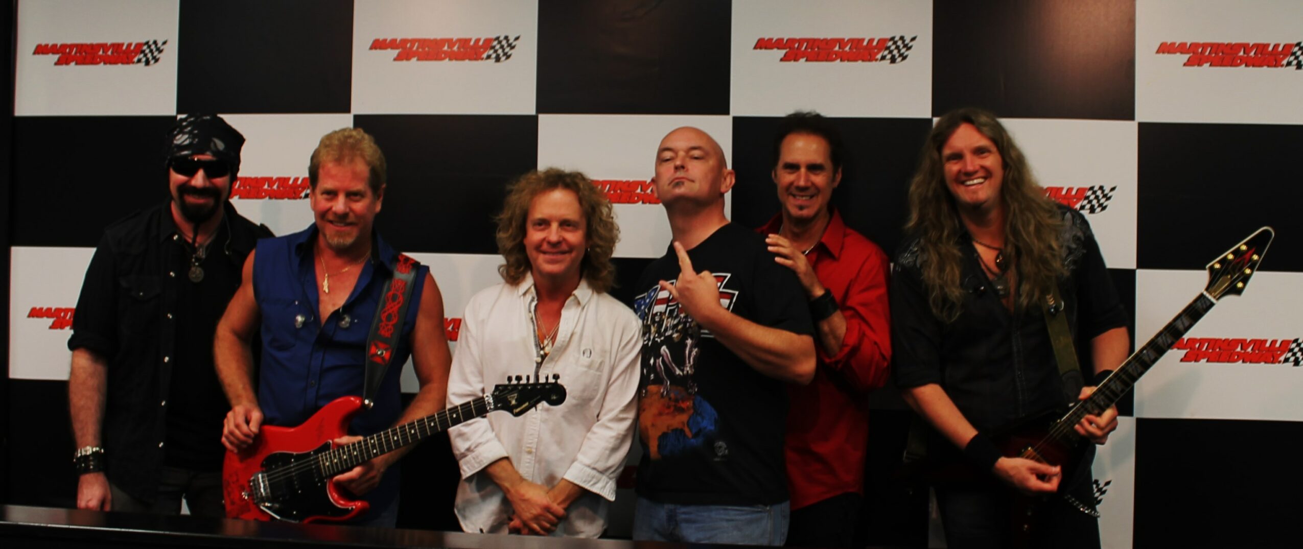 Six people standing in front of a checkered background, some holding guitars, smiling and posing for the photo.