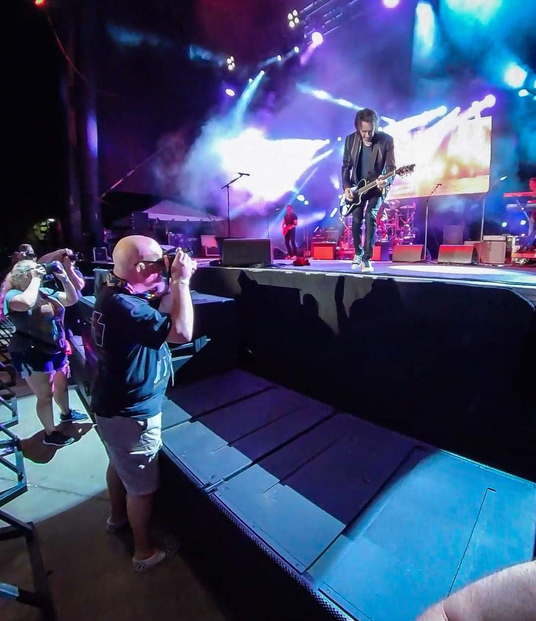Musician performing on stage with colorful lights, audience taking photos, and children watching nearby.