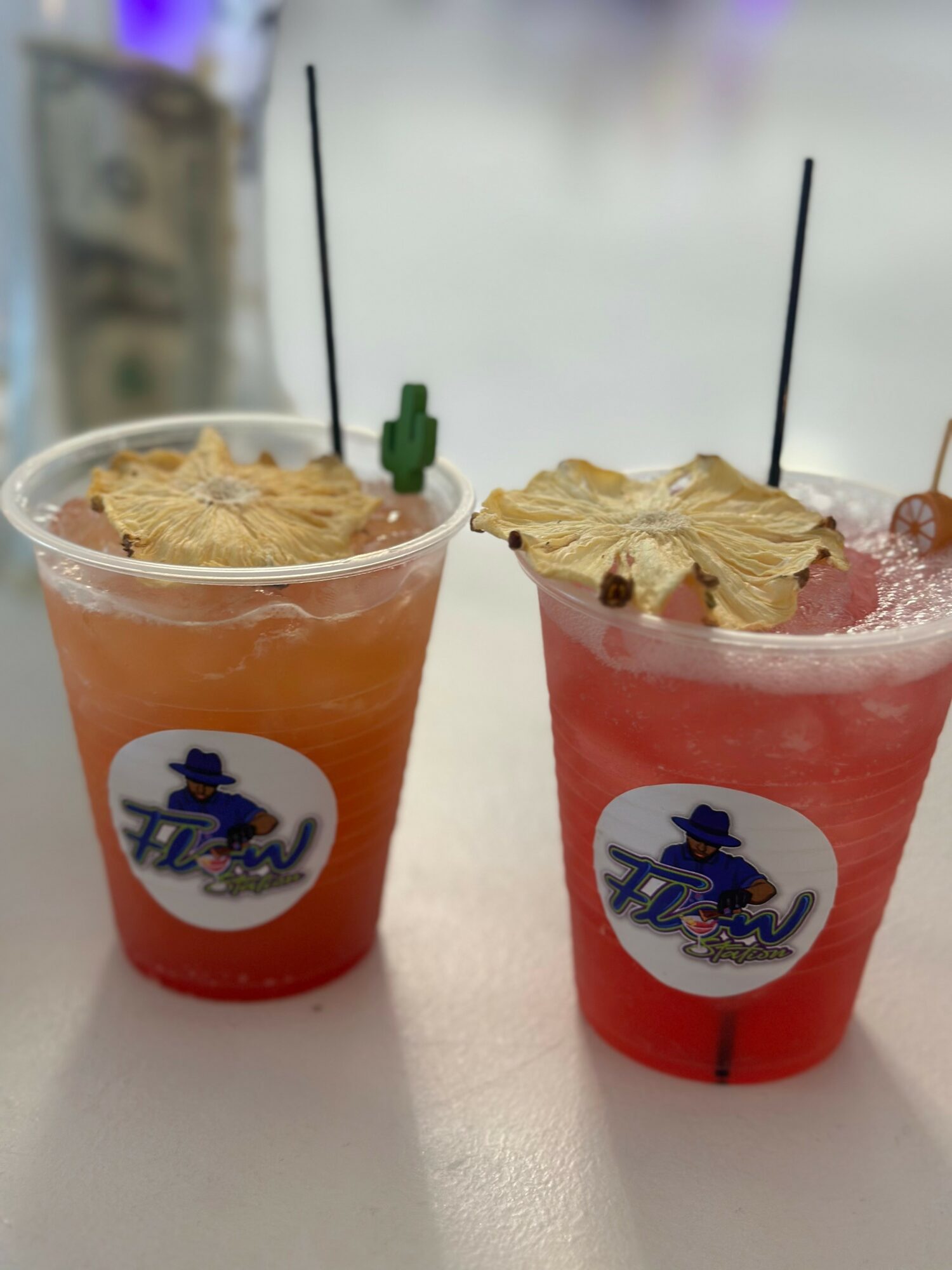 Two colorful drinks with whipped toppings and black straws on a white surface, with a blurred background.