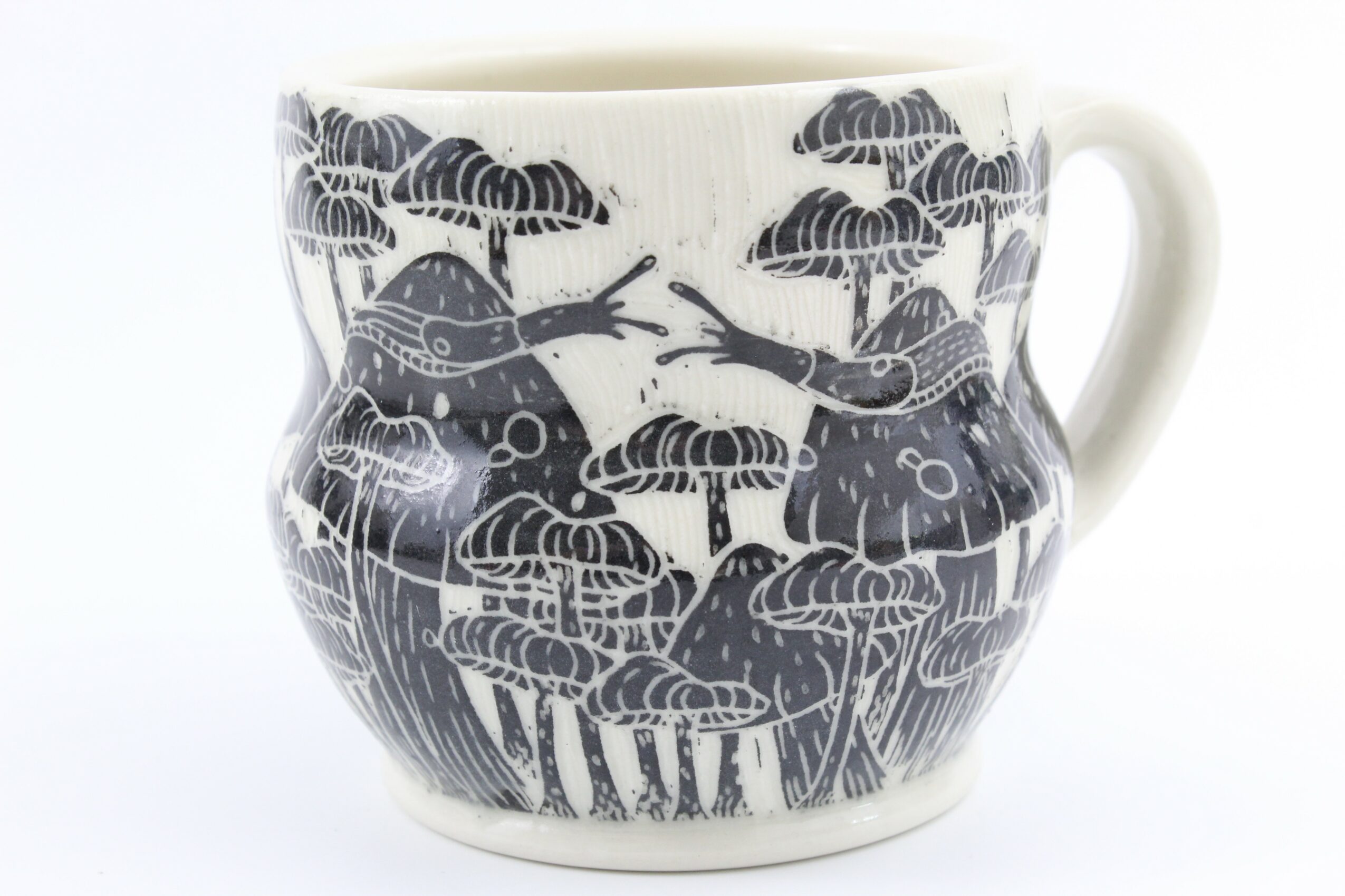 A white mug with a black and white illustration of trees and animals, including frogs and birds, in a natural scene.