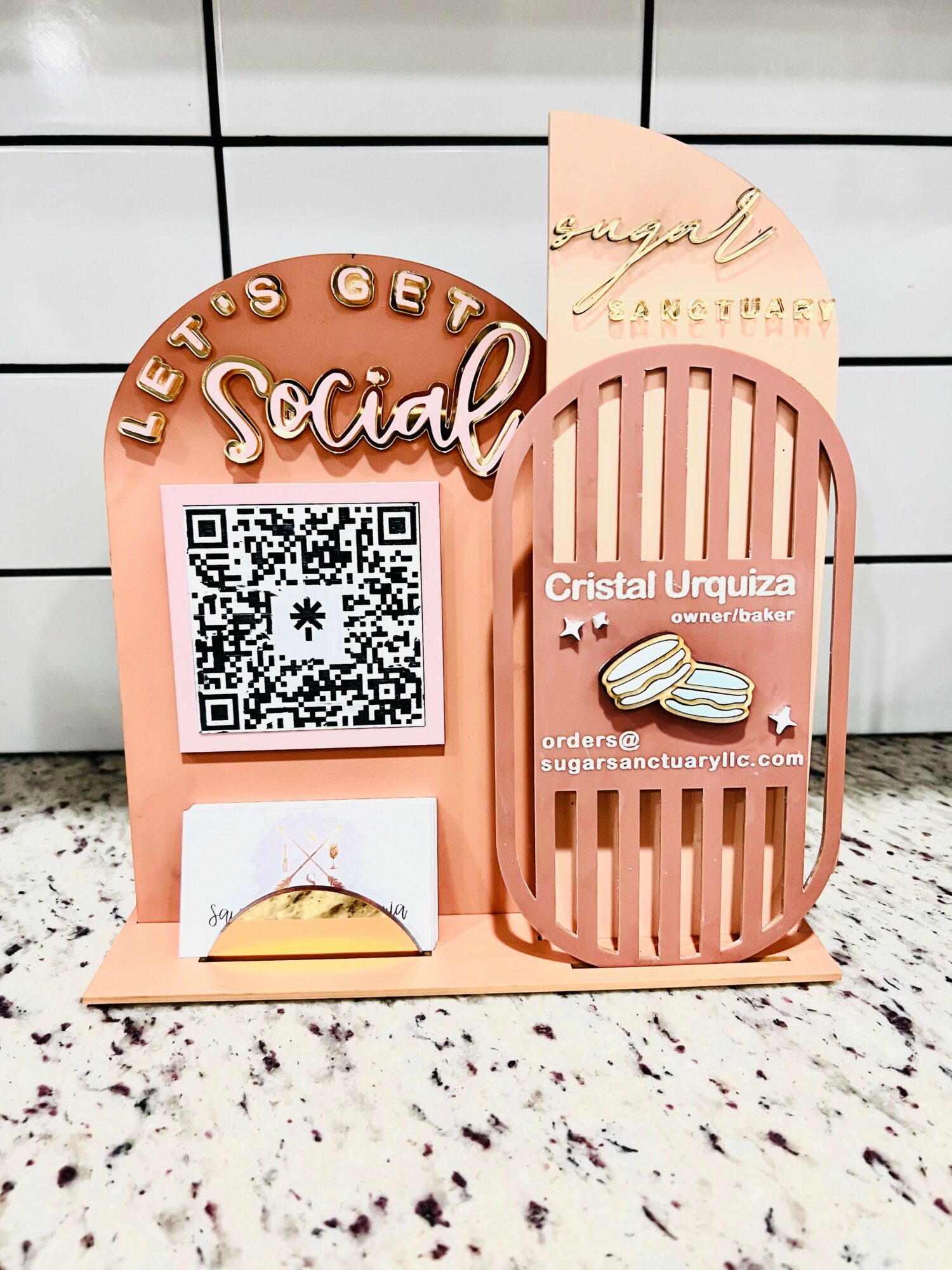 Pink social media stand with QR code, cookies, and decorative text, on a tiled surface. The stand has a circular sign and a slatted section.