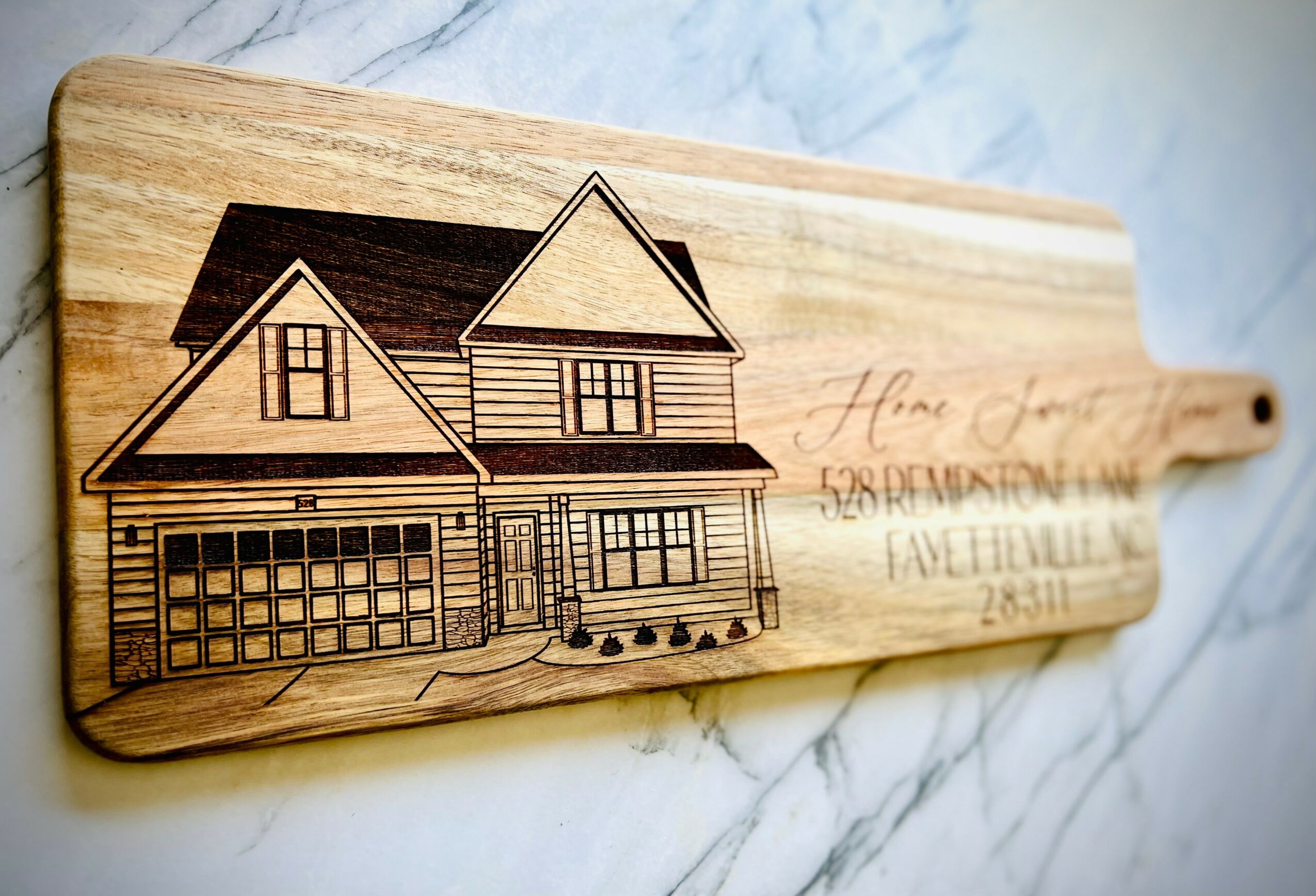 Wooden cutting board with house illustration and handwritten text, placed on a marble surface.