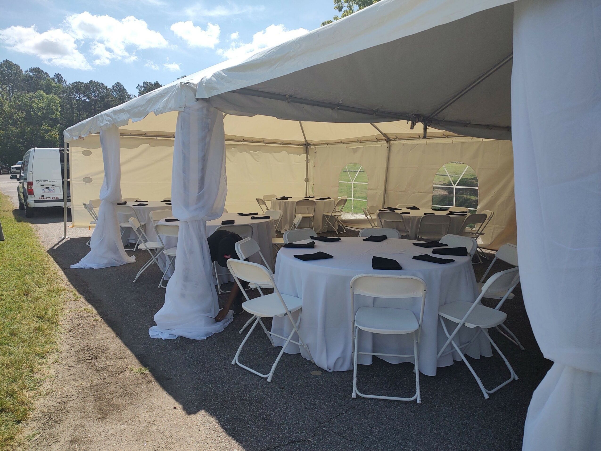 Event setup with a long table, white chairs, and tall white covered pillars under a tent, outdoors with a cloudy sky.