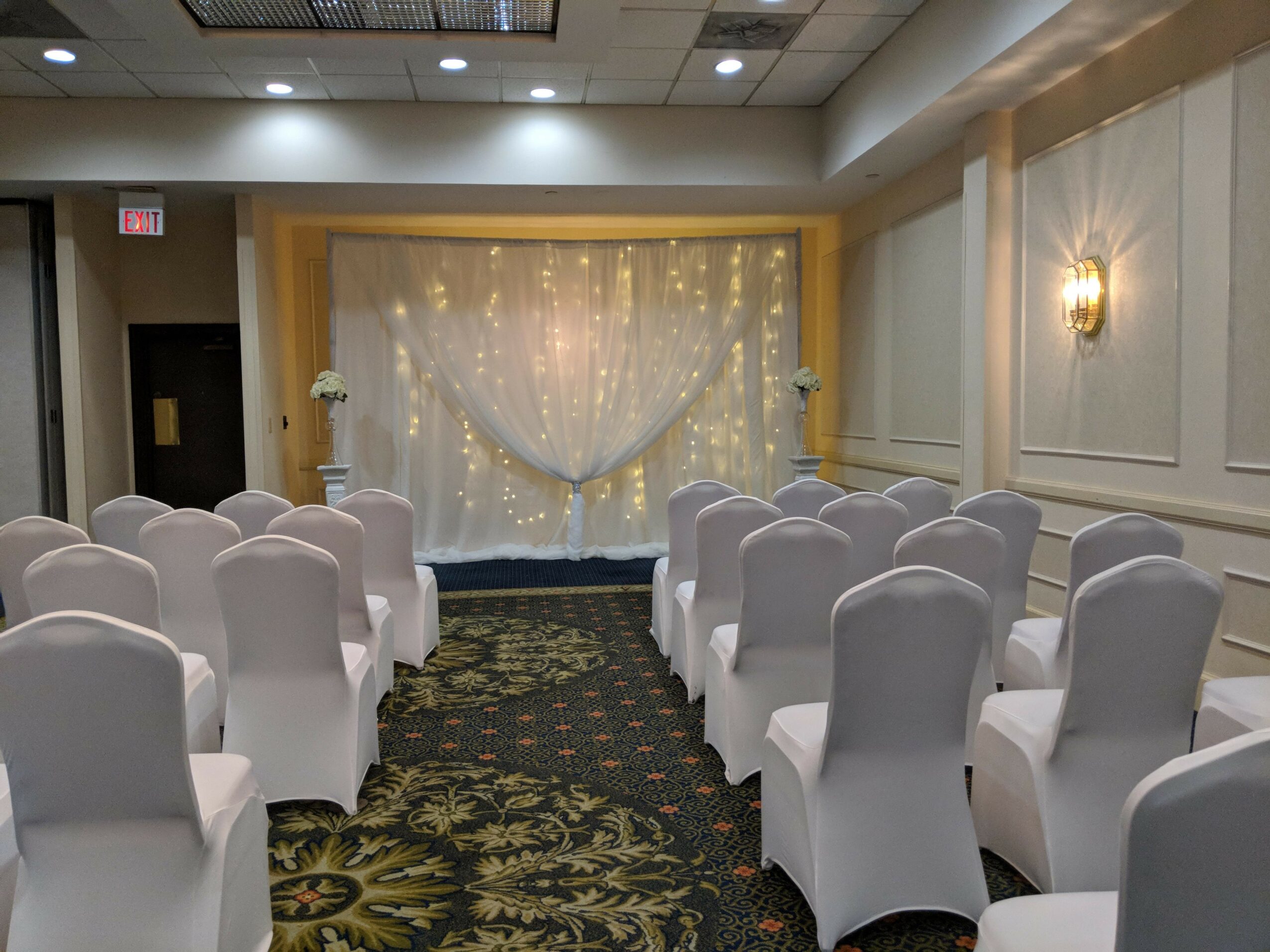 Empty event room with chairs facing decorated backdrop, floral arrangements, and string lights, ready for a ceremony or celebration.