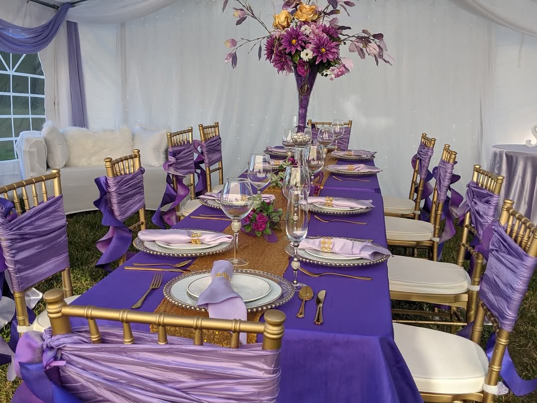 Long dining table with purple tablecloth, gold chairs with purple sashes, floral centerpiece, and place settings inside a decorated tent.