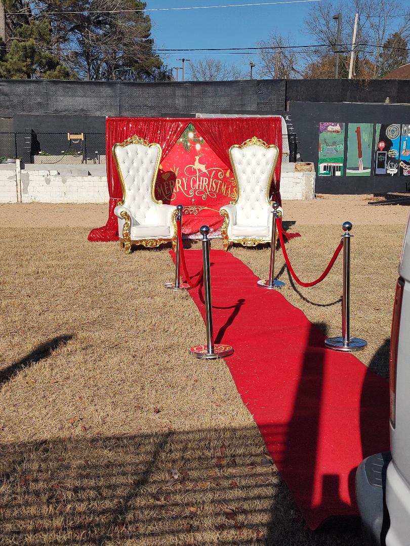 Two ornate white chairs with gold accents on a red carpet, with a backdrop reading 'Merry Christmas,' outdoors on grass.